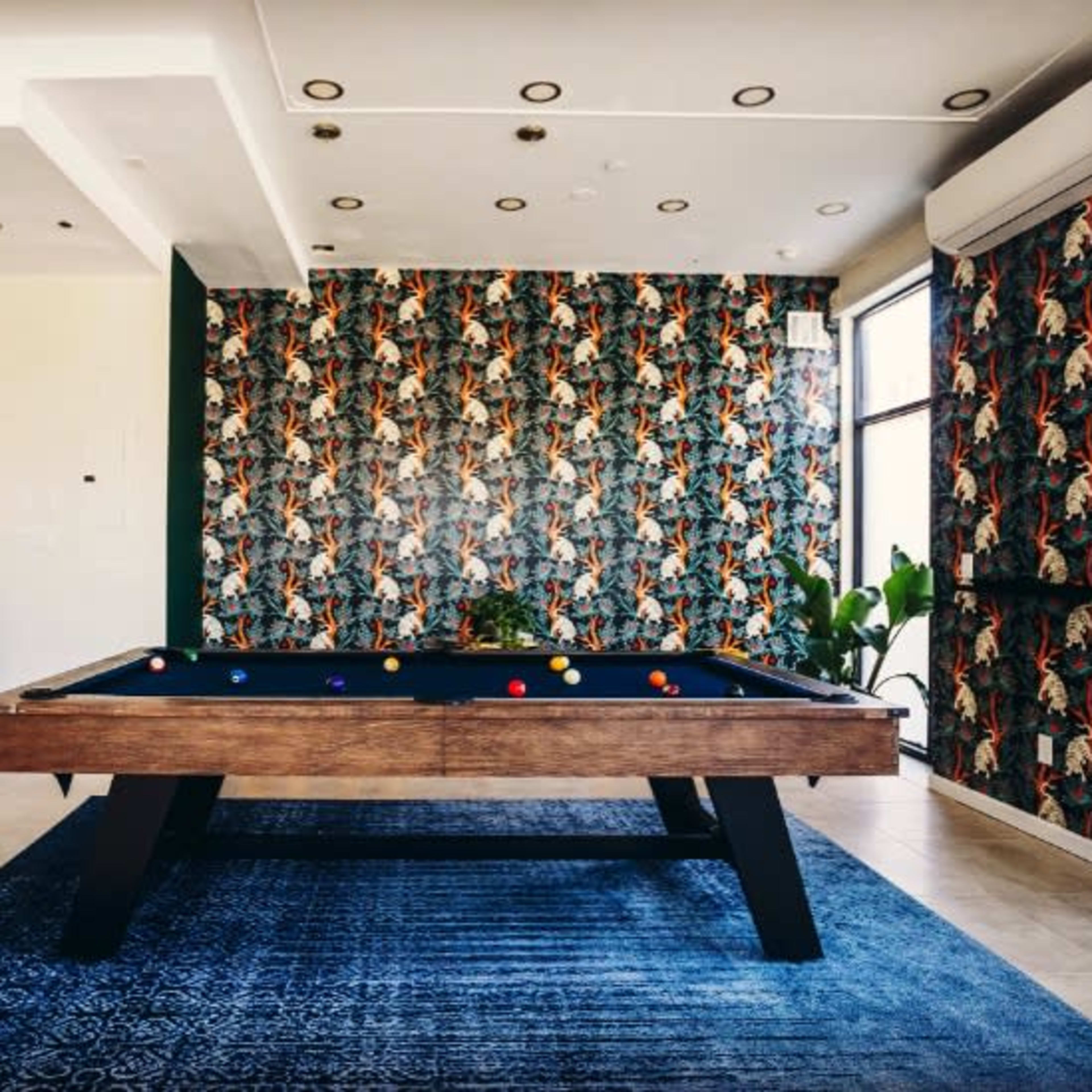 A modern game room featuring a billiards table, patterned wallpaper, and large windows with plants nearby.