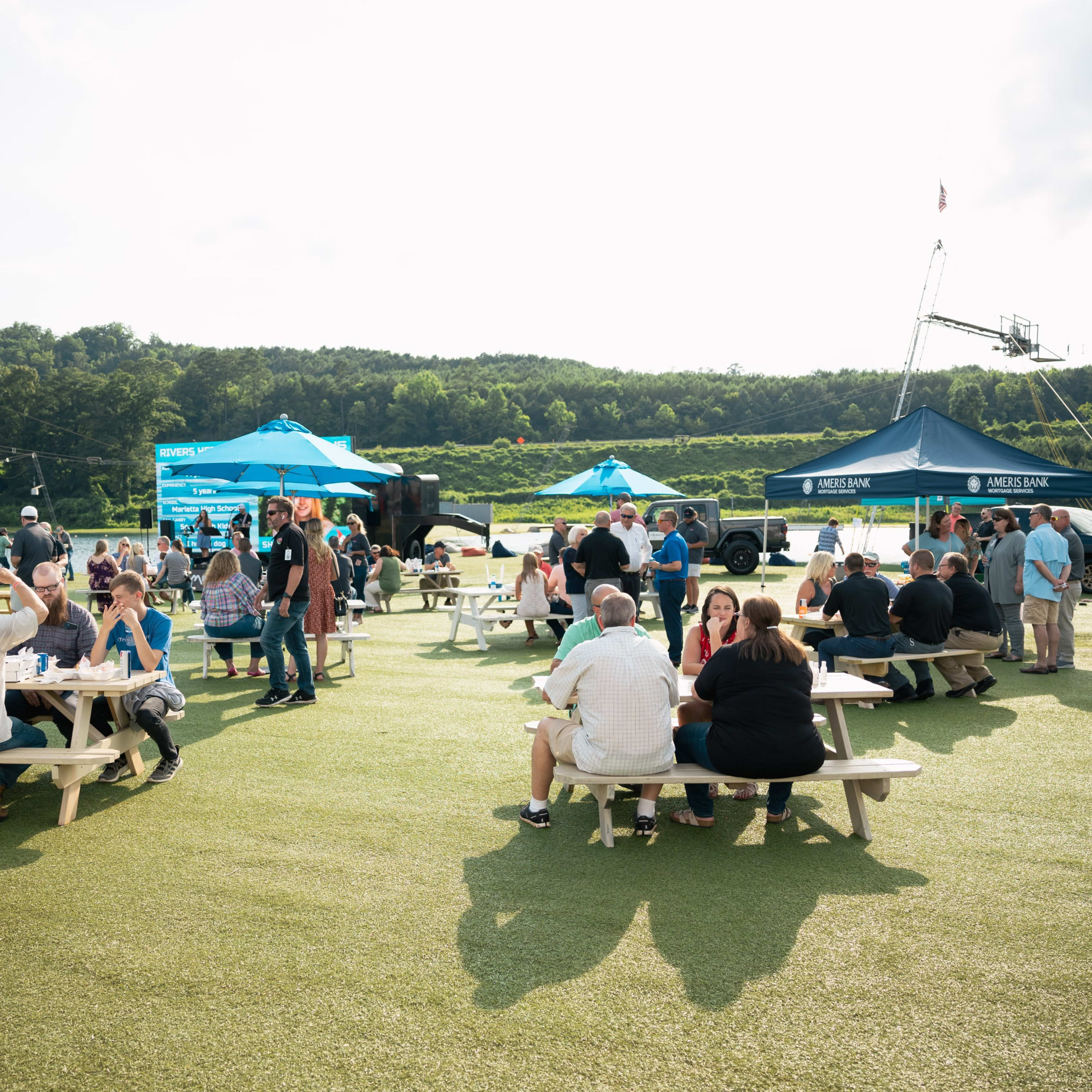 A large group of people socializes around picnic tables at an outdoor event on a green field, with tents and a scenic background.
