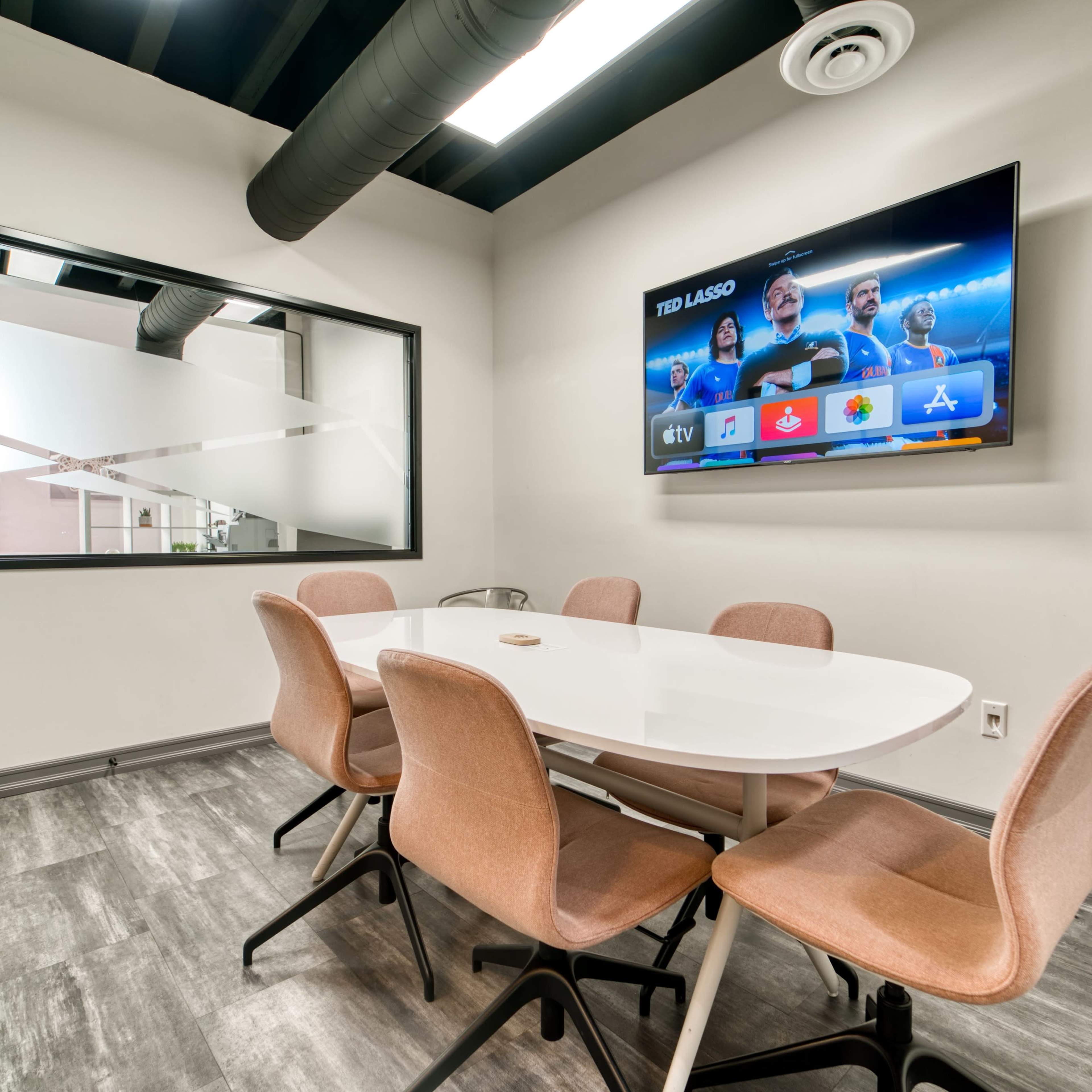 A modern conference room with a round white table, six beige chairs, a wall-mounted TV displaying a screen, and a landline phone on a side table.