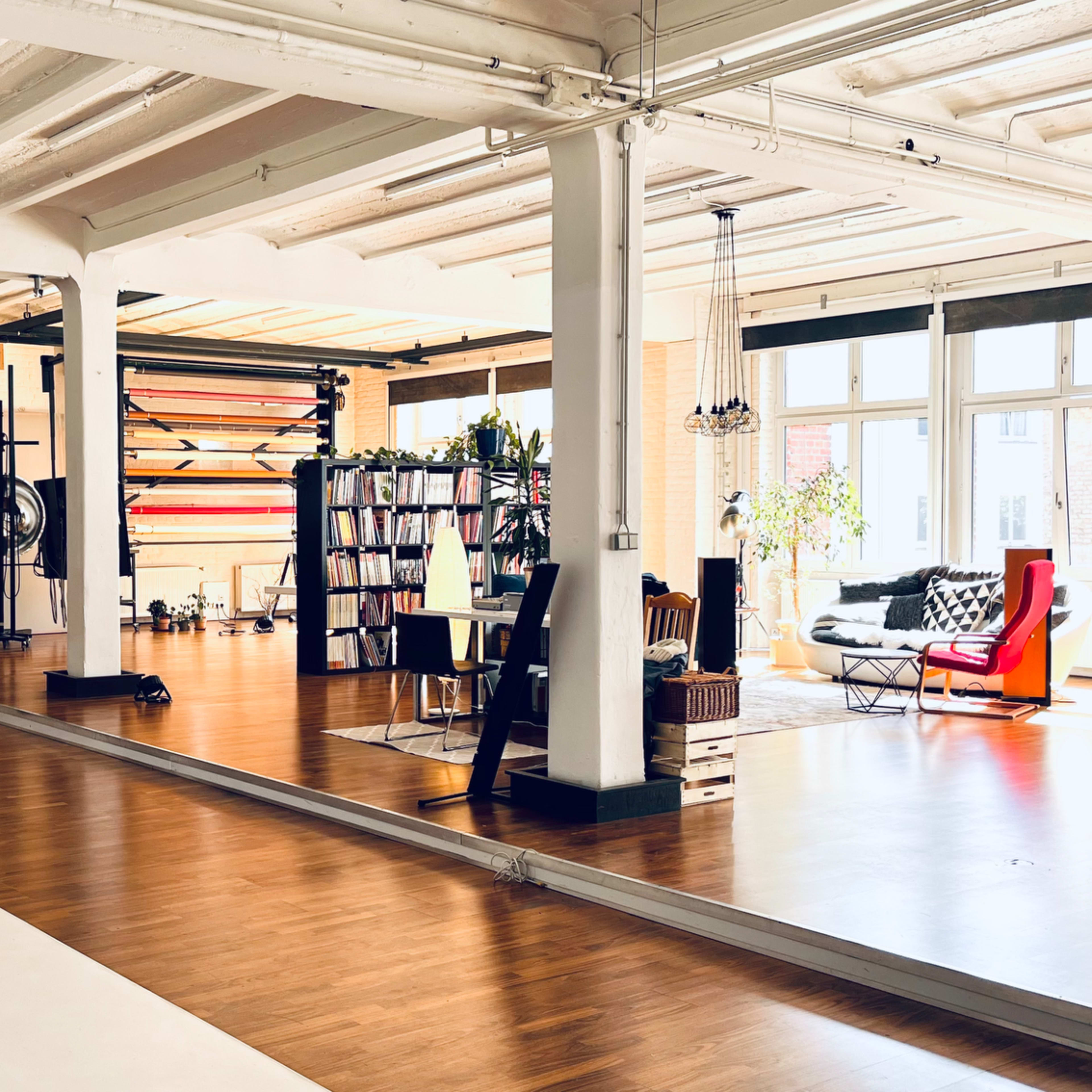 A spacious, well-lit studio featuring wooden floors, large windows, and various furniture arrangements, including a red chair and bookshelves.