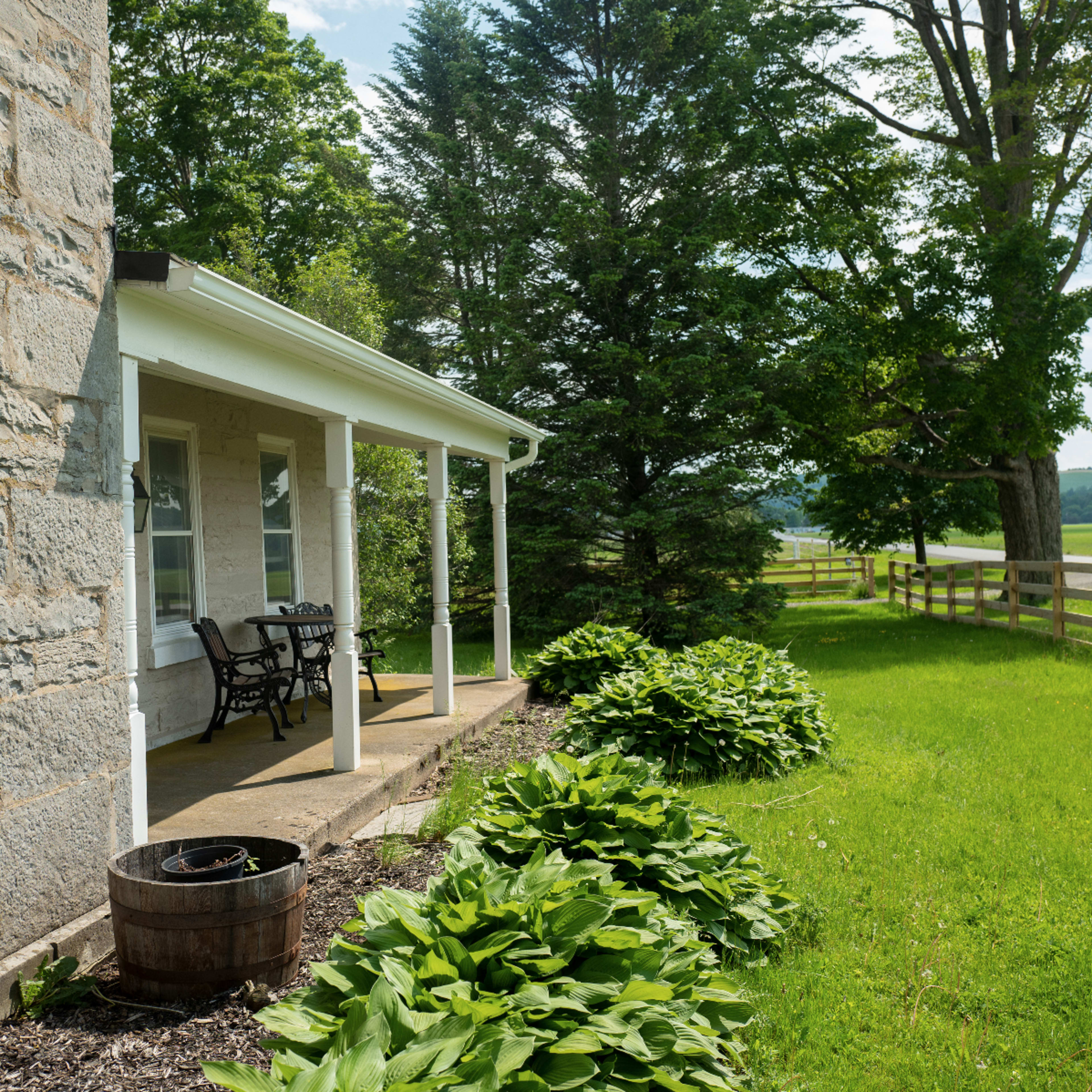 A stone house with a porch, surrounded by a grassy yard and a wooden fence in the background.