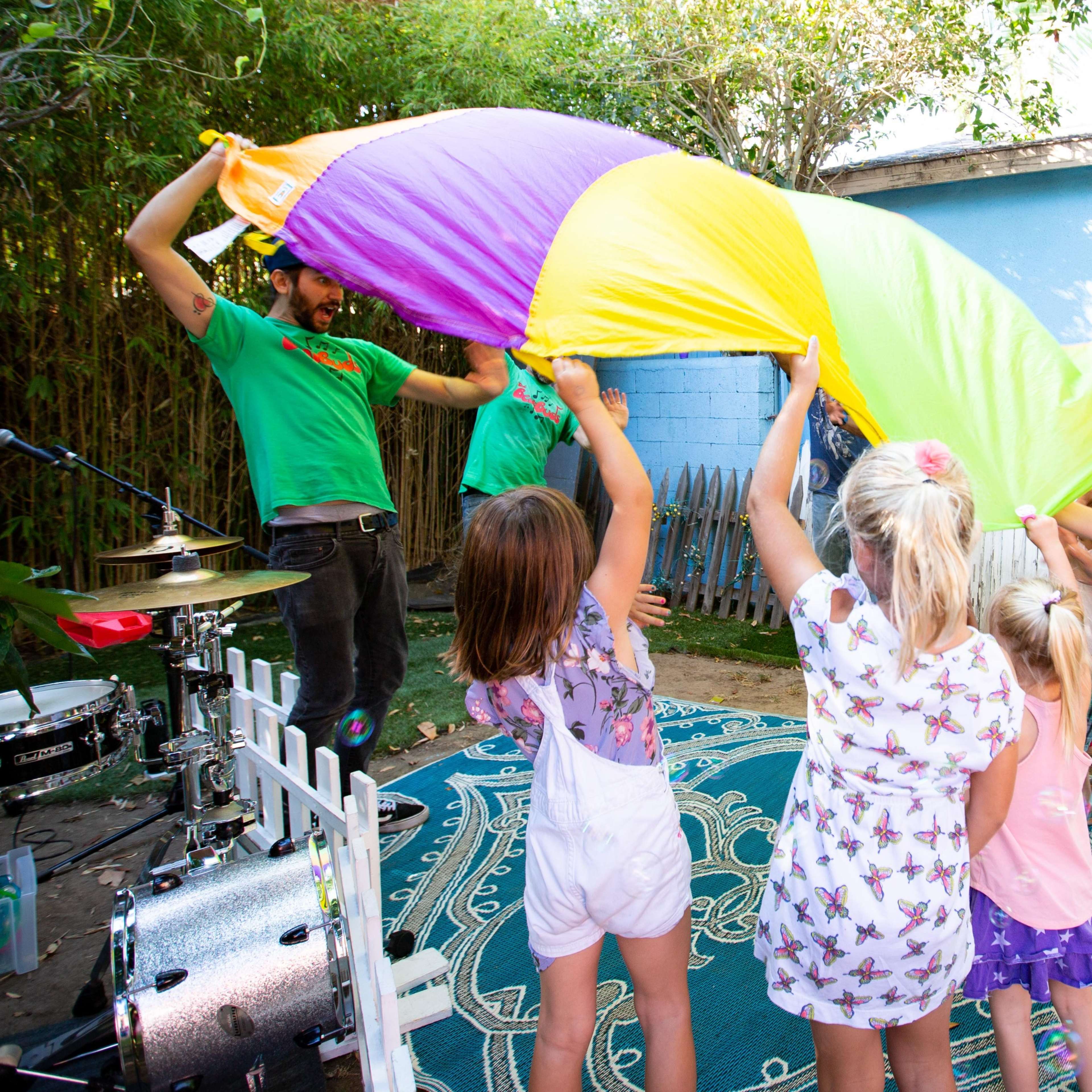 Children are playing with a large purple and yellow parachute while an adult musician performs nearby.
