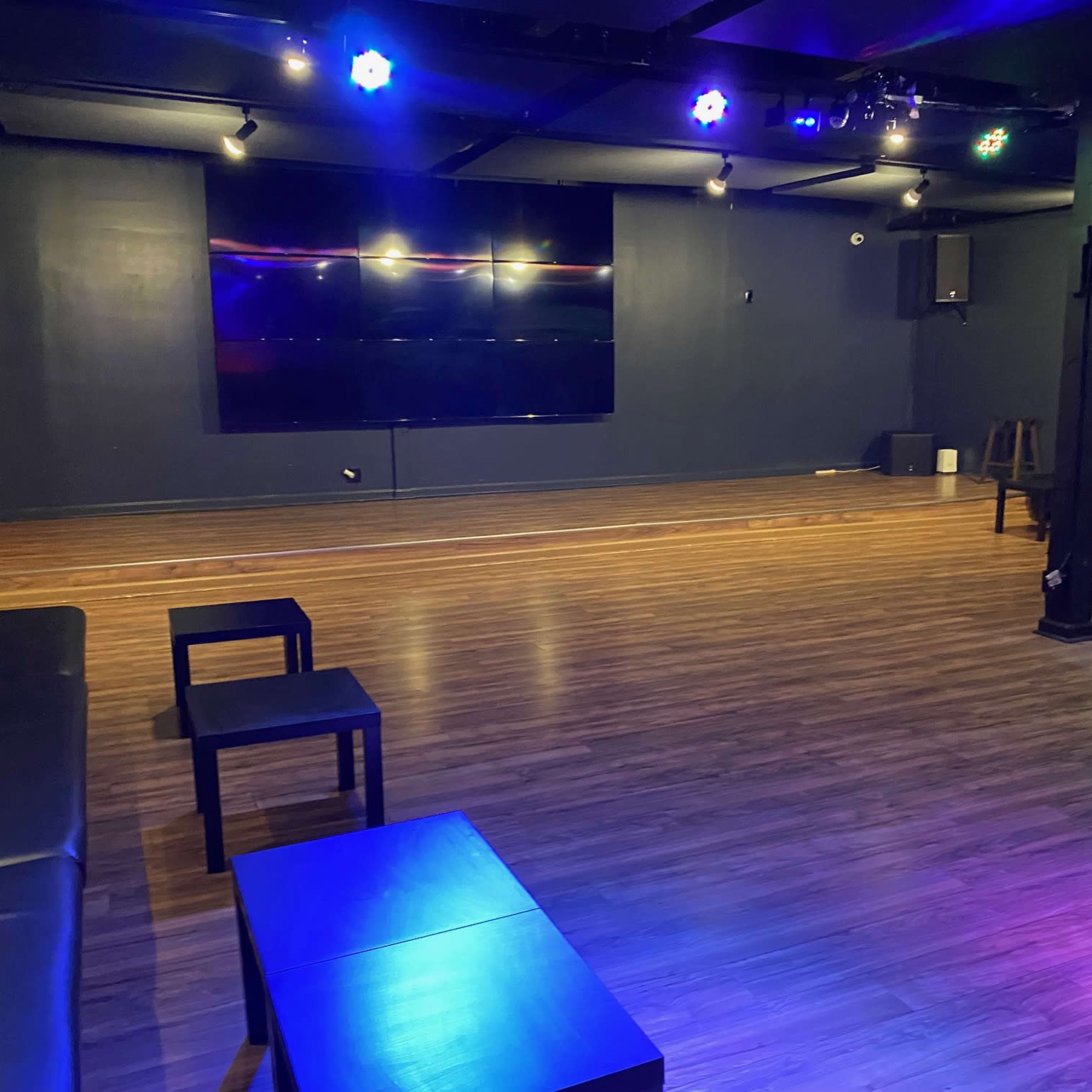 A spacious, empty room with a wooden floor, black seating, and a large screen on the wall, illuminated by multicolored lights.