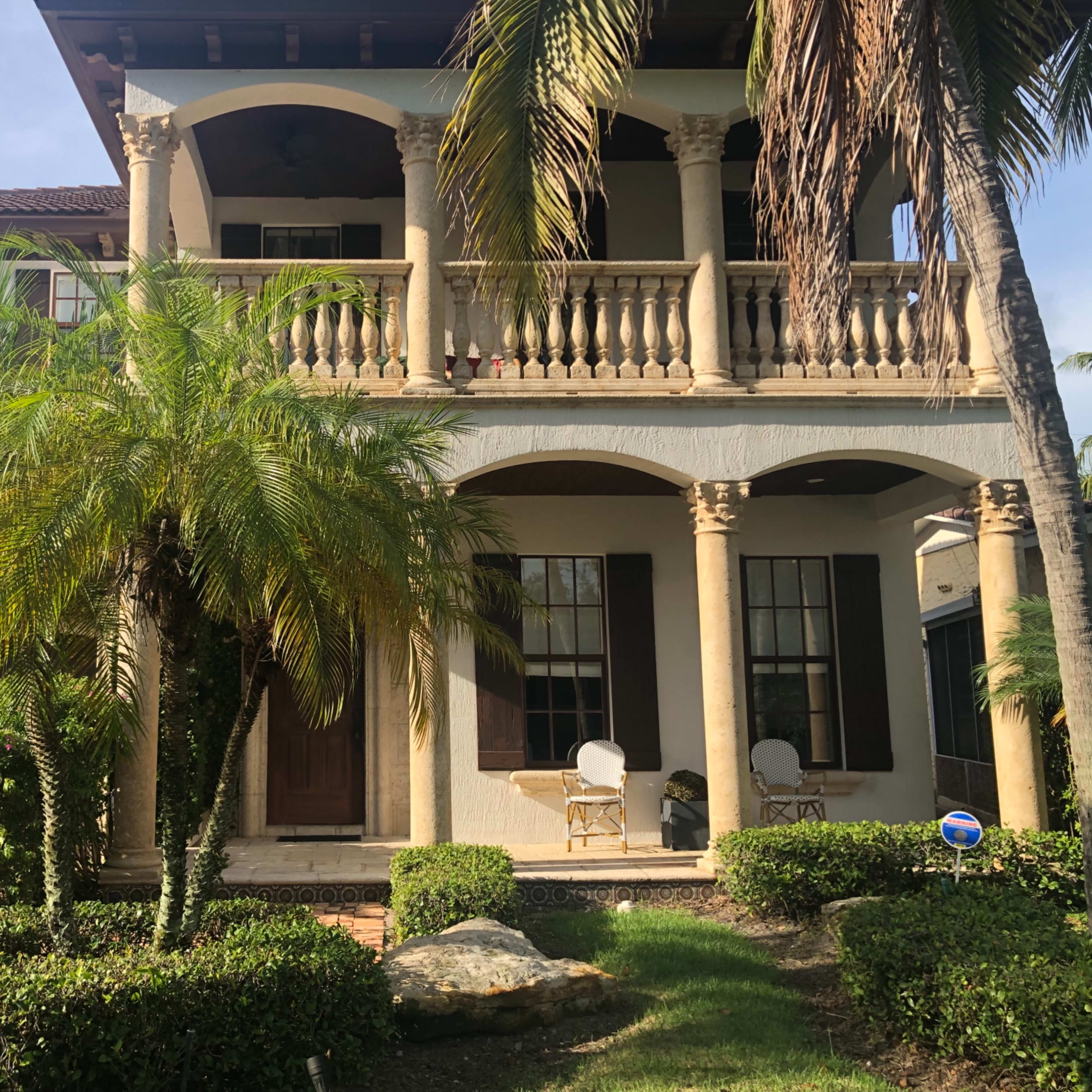 A two-story house features a large front porch with columns, surrounded by palm trees and neatly trimmed hedges.
