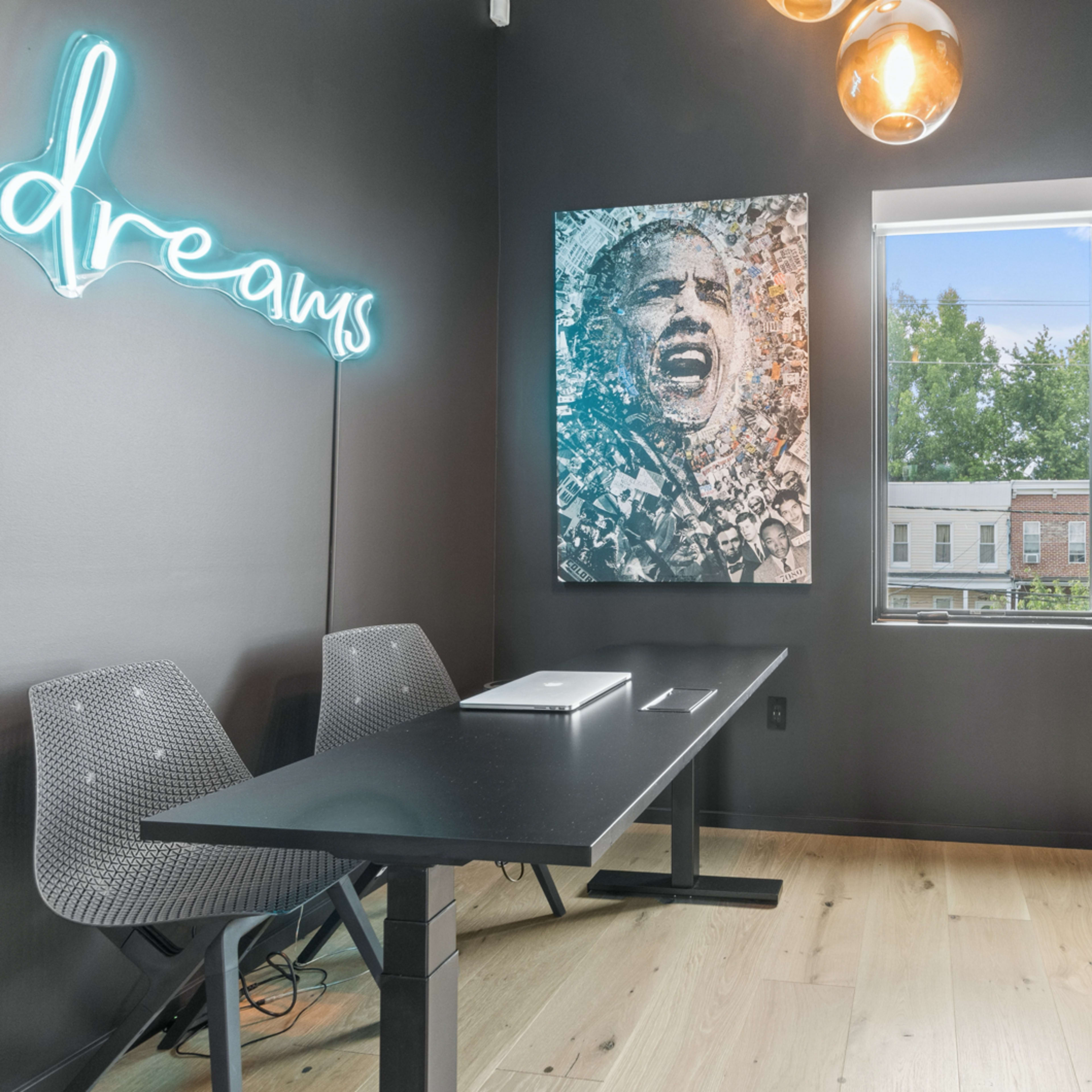 A modern workspace features a black wall with a large portrait of a man, a desk with a laptop, and a window providing natural light.