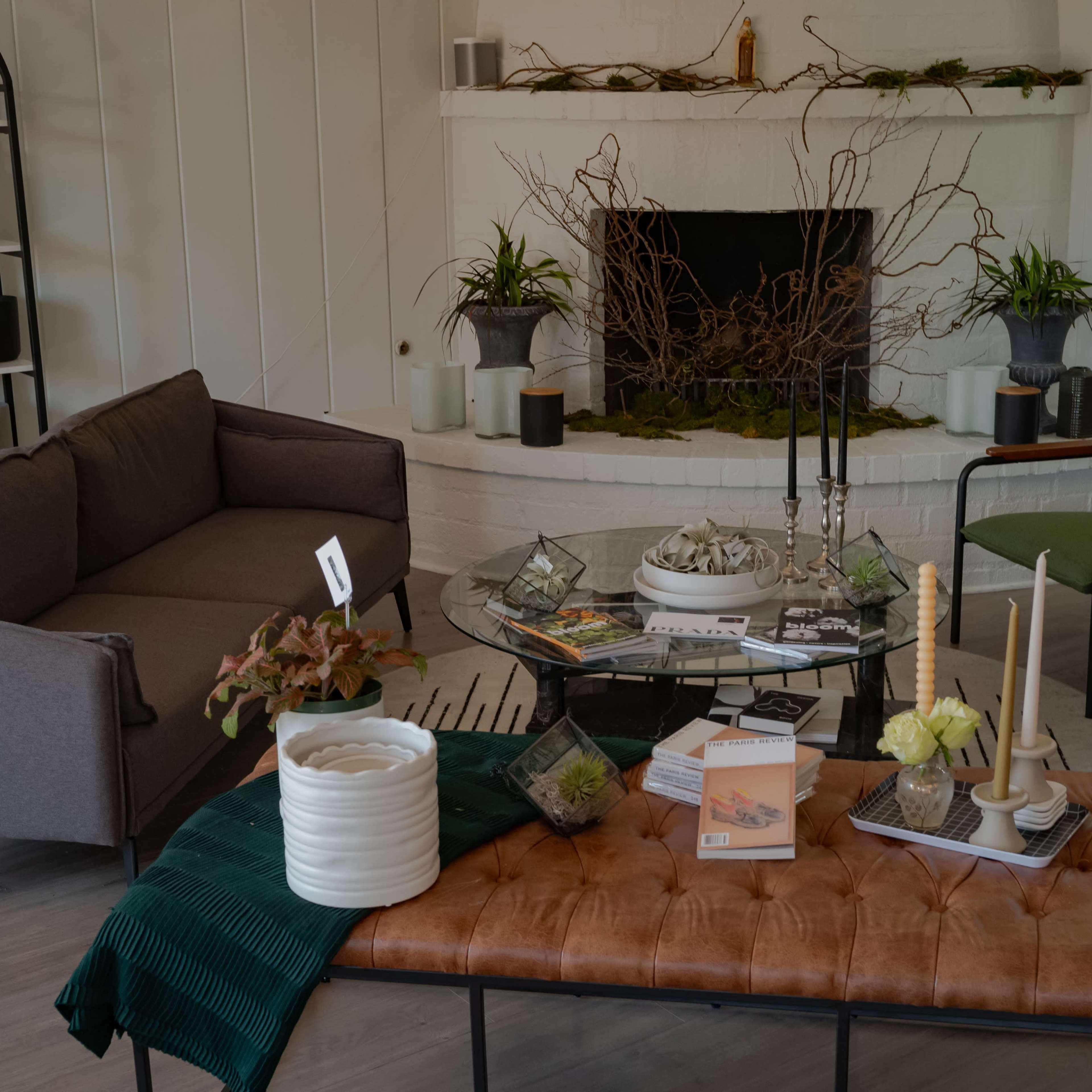 A cozy living room features a gray sofa, two green chairs, a coffee table with decorative items, and a fireplace adorned with plants and branches.