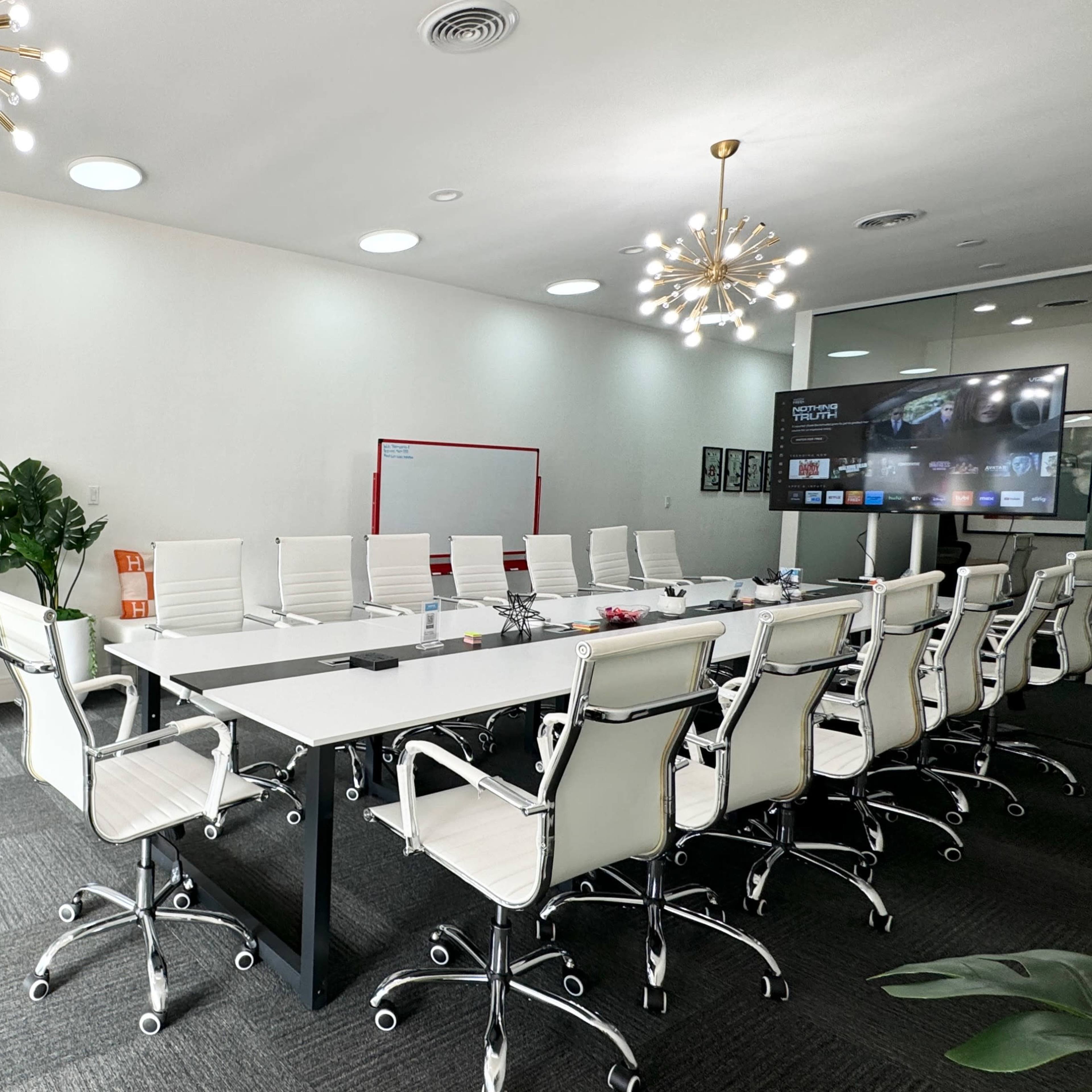 A modern conference room features a long table surrounded by white chairs, a large screen on one wall, and decorative elements including plants and a framed picture.