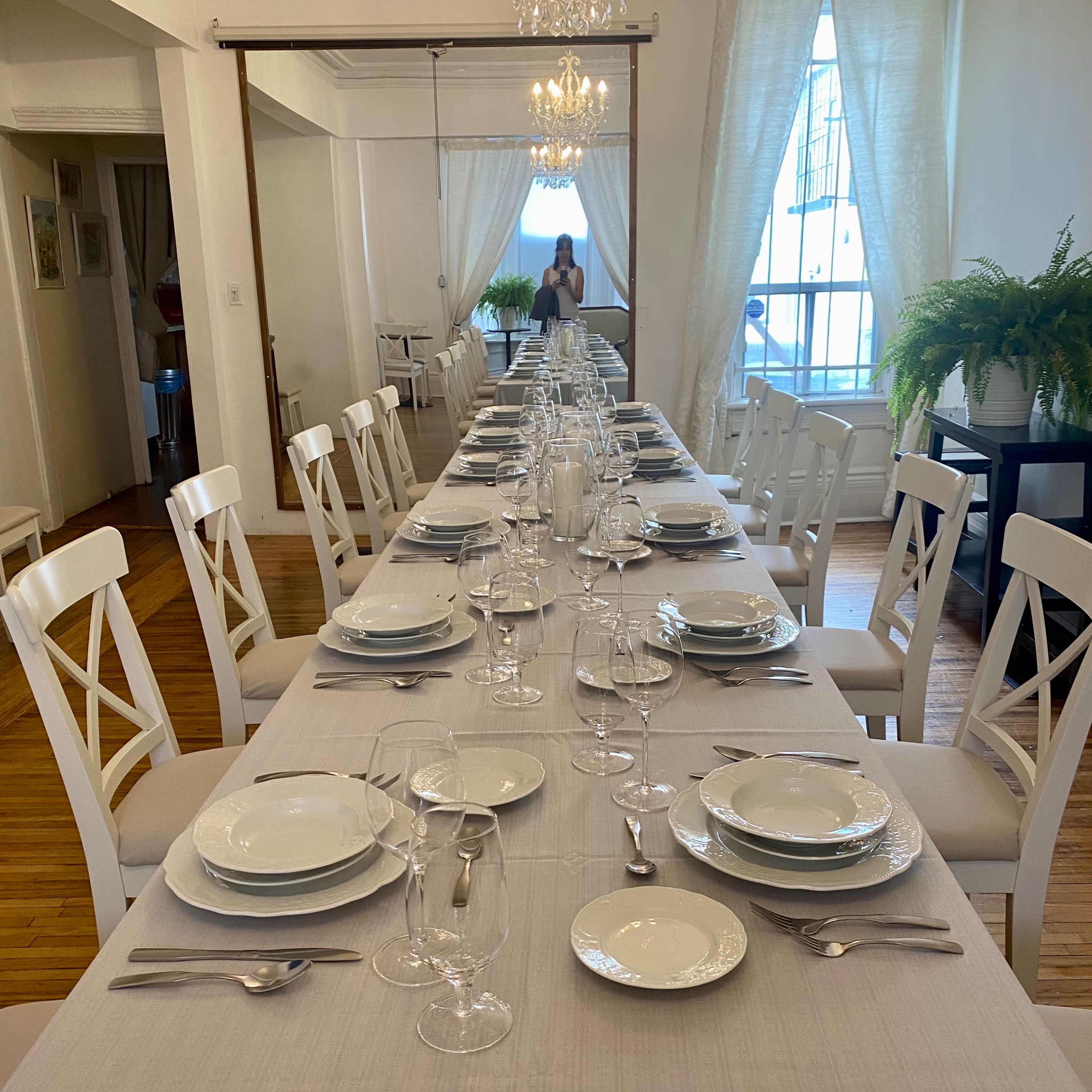A long dining table is set with plates, glasses, and cutlery under a chandelier in a well-lit room.