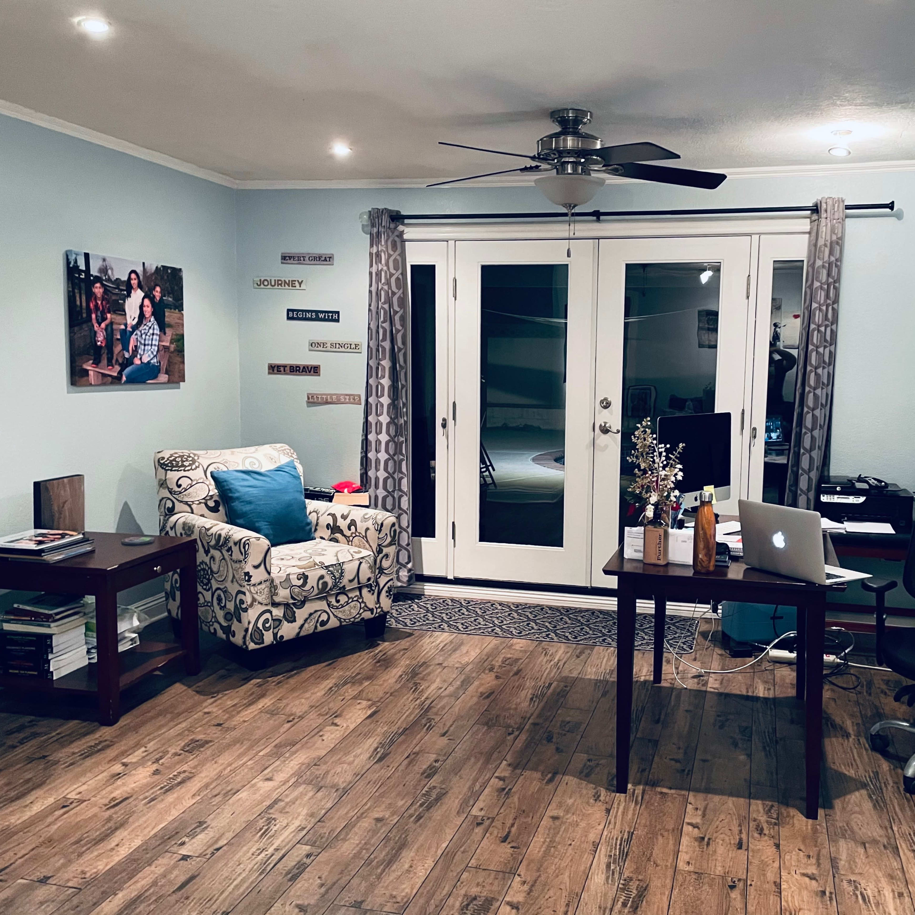 A cozy room features a patterned armchair, a wooden table with a laptop, and a workspace with a black office chair, alongside glass doors leading outside.
