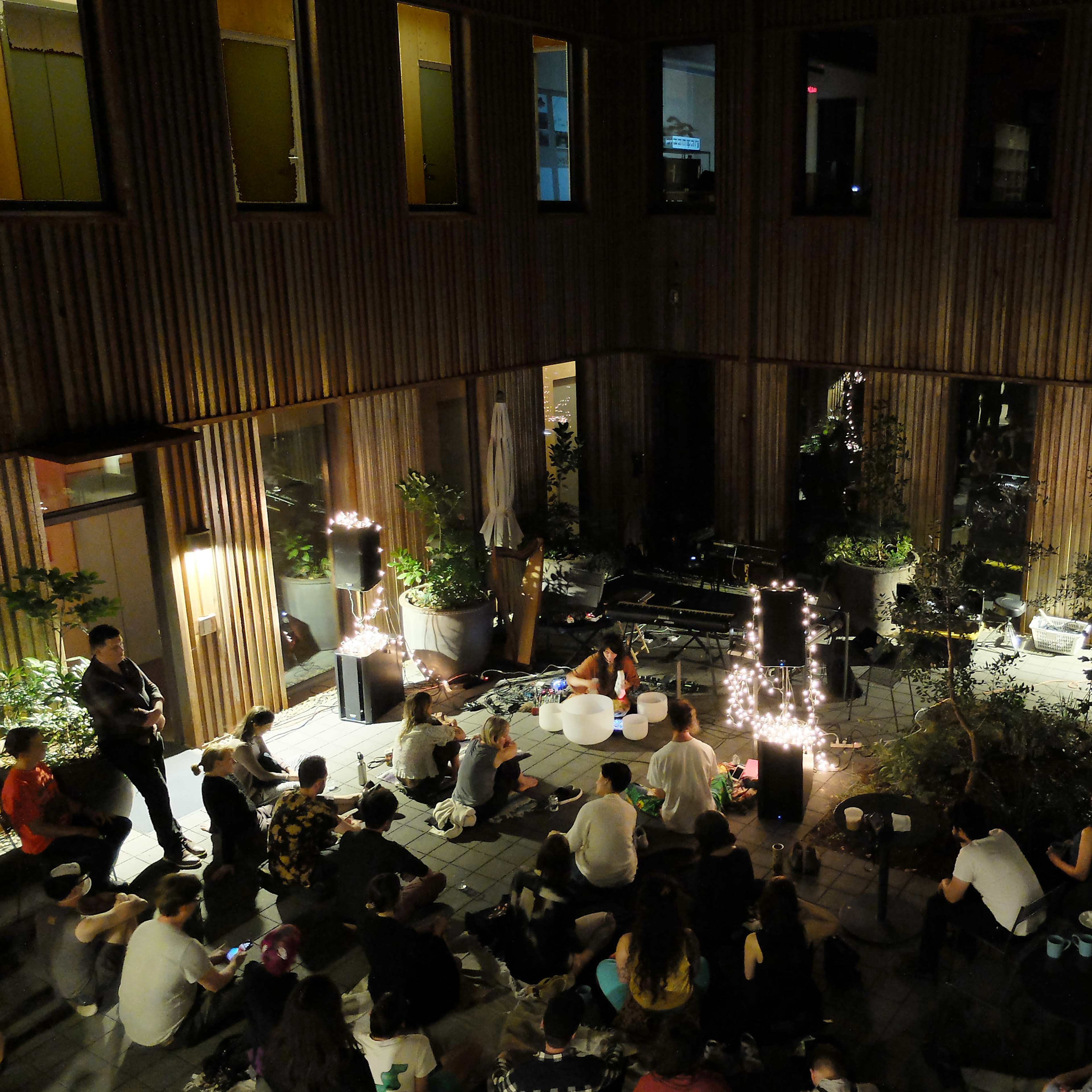 A group of people is seated in a courtyard surrounded by wooden walls, listening to a performer near a piano and decorative lights.