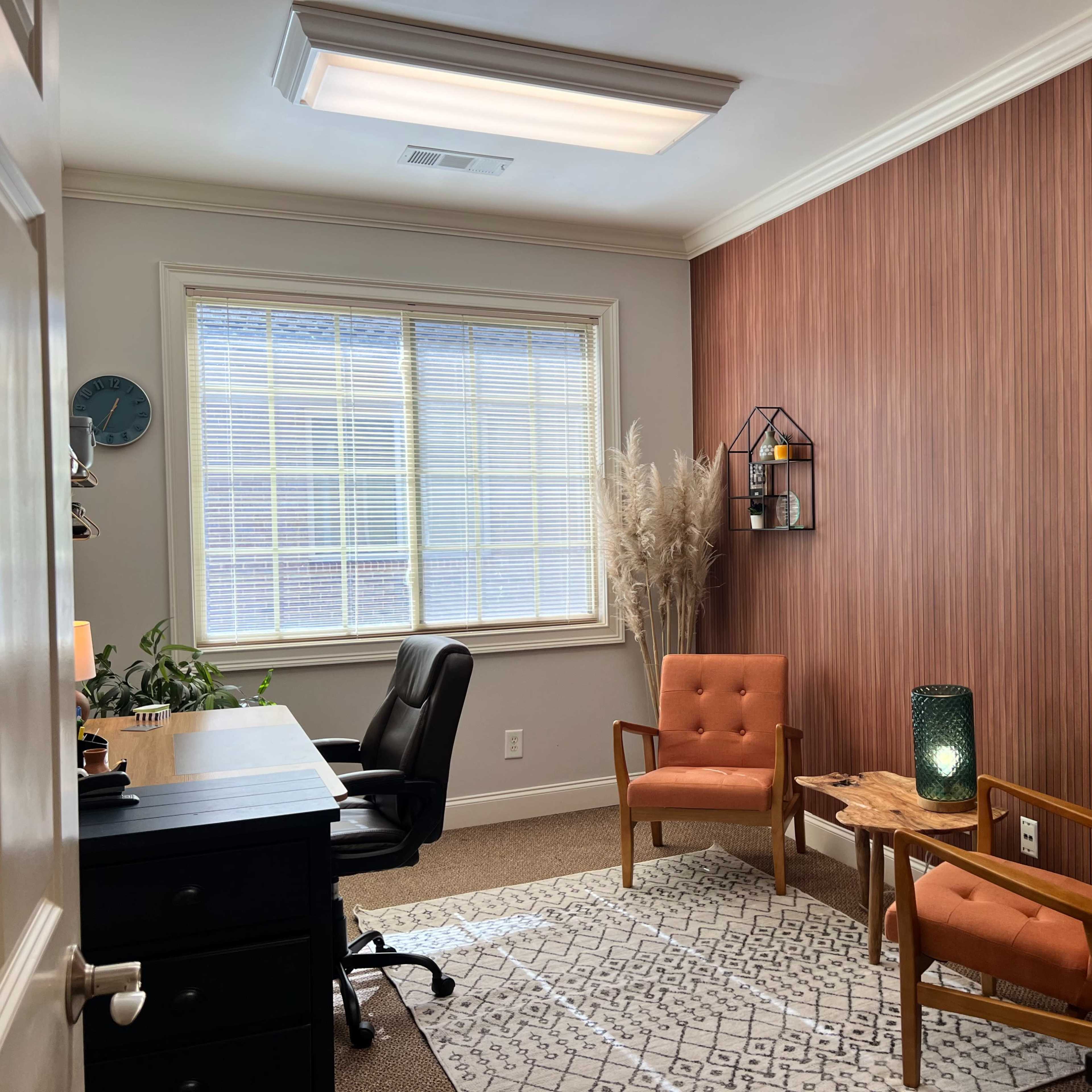 A well-lit office space featuring two orange chairs, a desk with a black chair, and decorative elements including a plant and wall hangings.