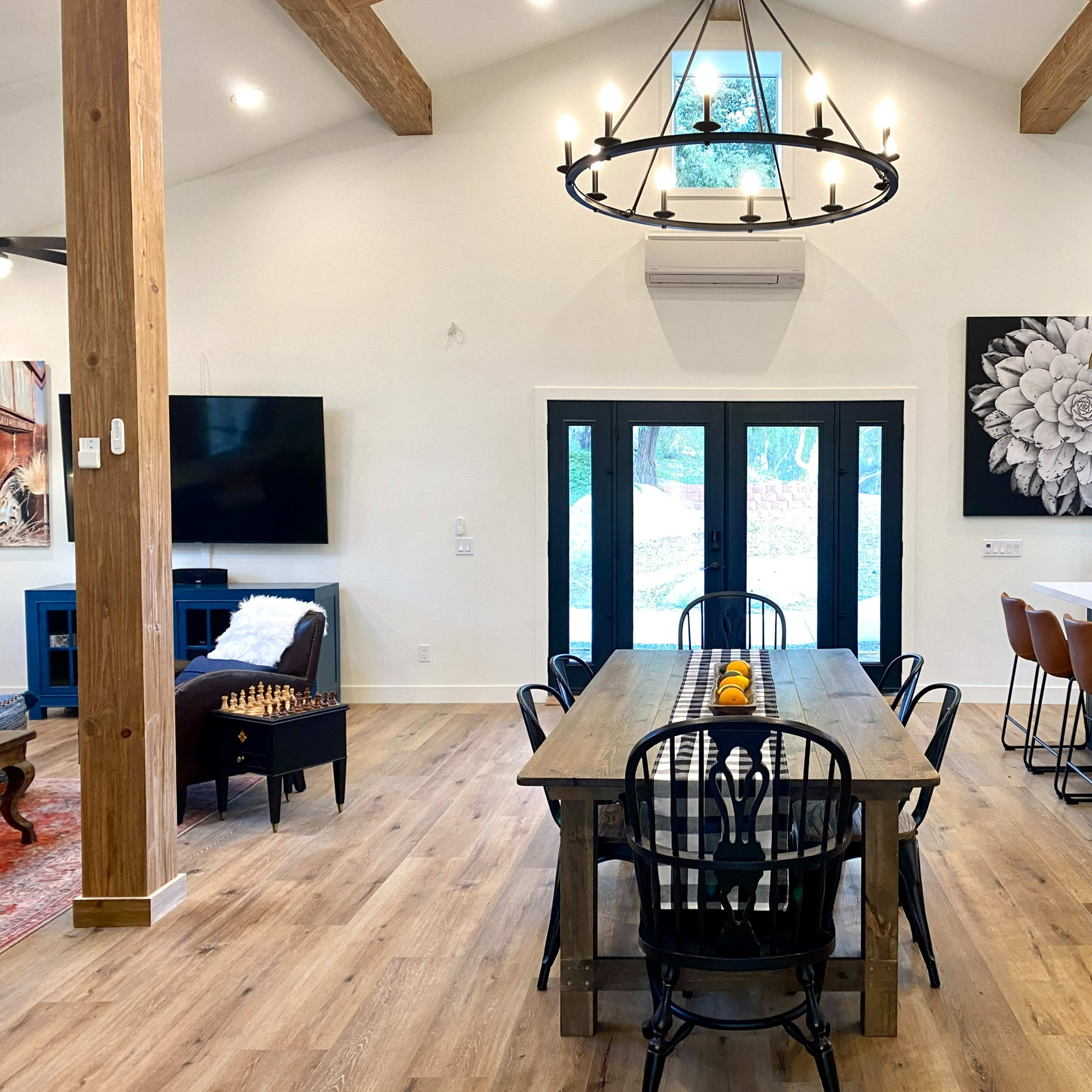 A spacious dining area features a long wooden table with black chairs, a chandelier overhead, and large windows overlooking an outdoor view.