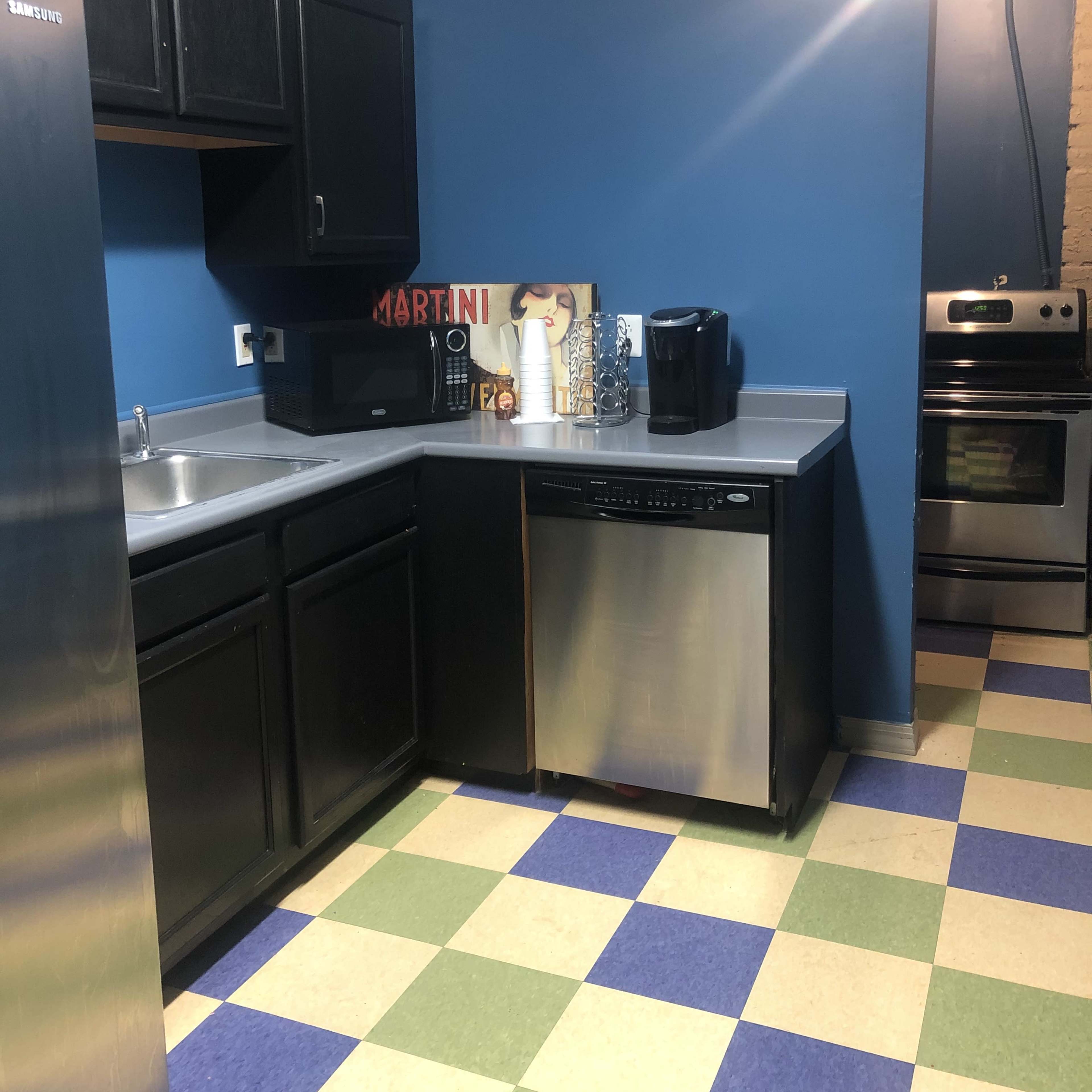 A kitchen with dark cabinets, a stainless steel dishwasher, a microwave, and a coffee maker on a countertop, set against a blue wall and checkered green and blue floor tiles.