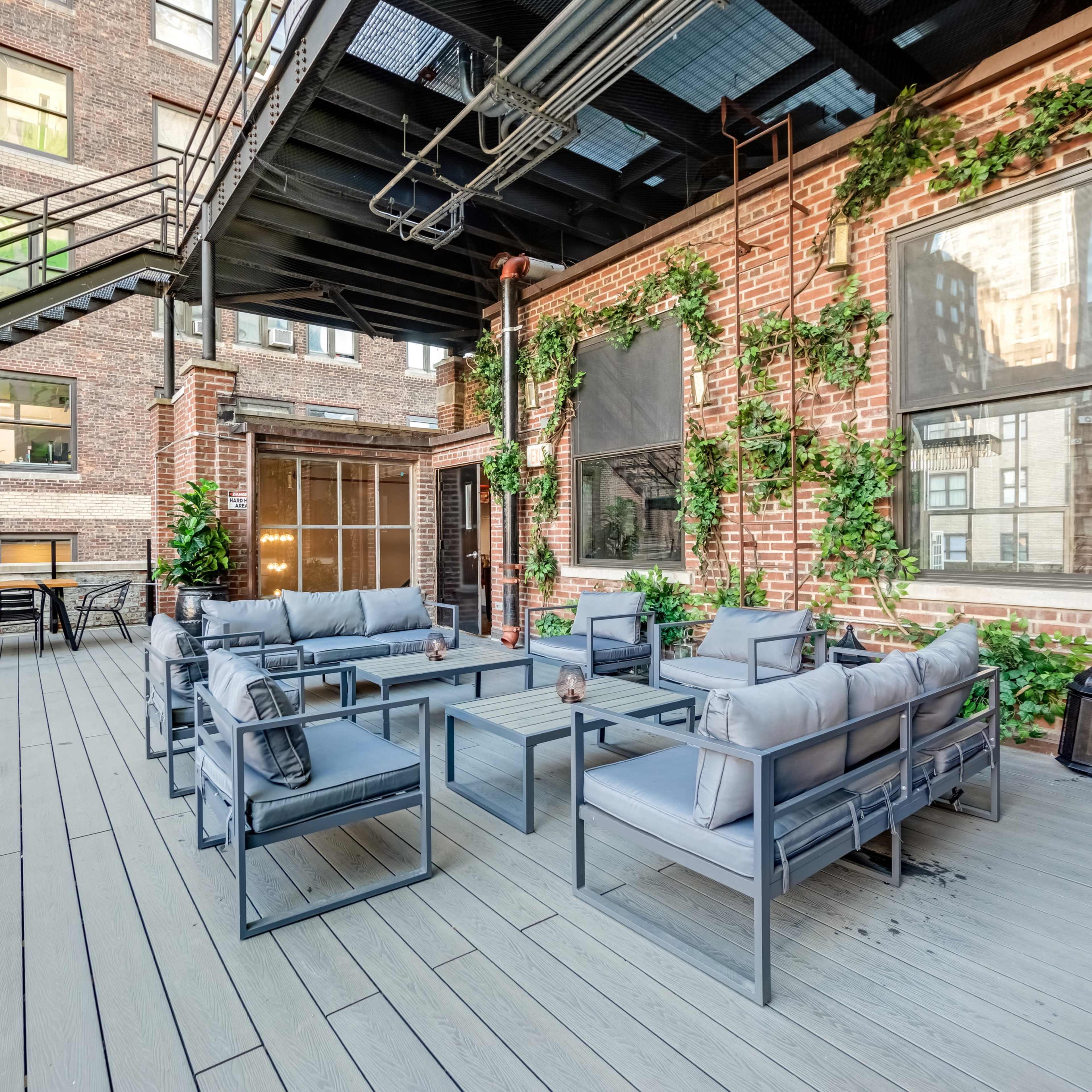 An outdoor rooftop terrace with seating areas, surrounded by brick walls and greenery, featuring a staircase and large windows.
