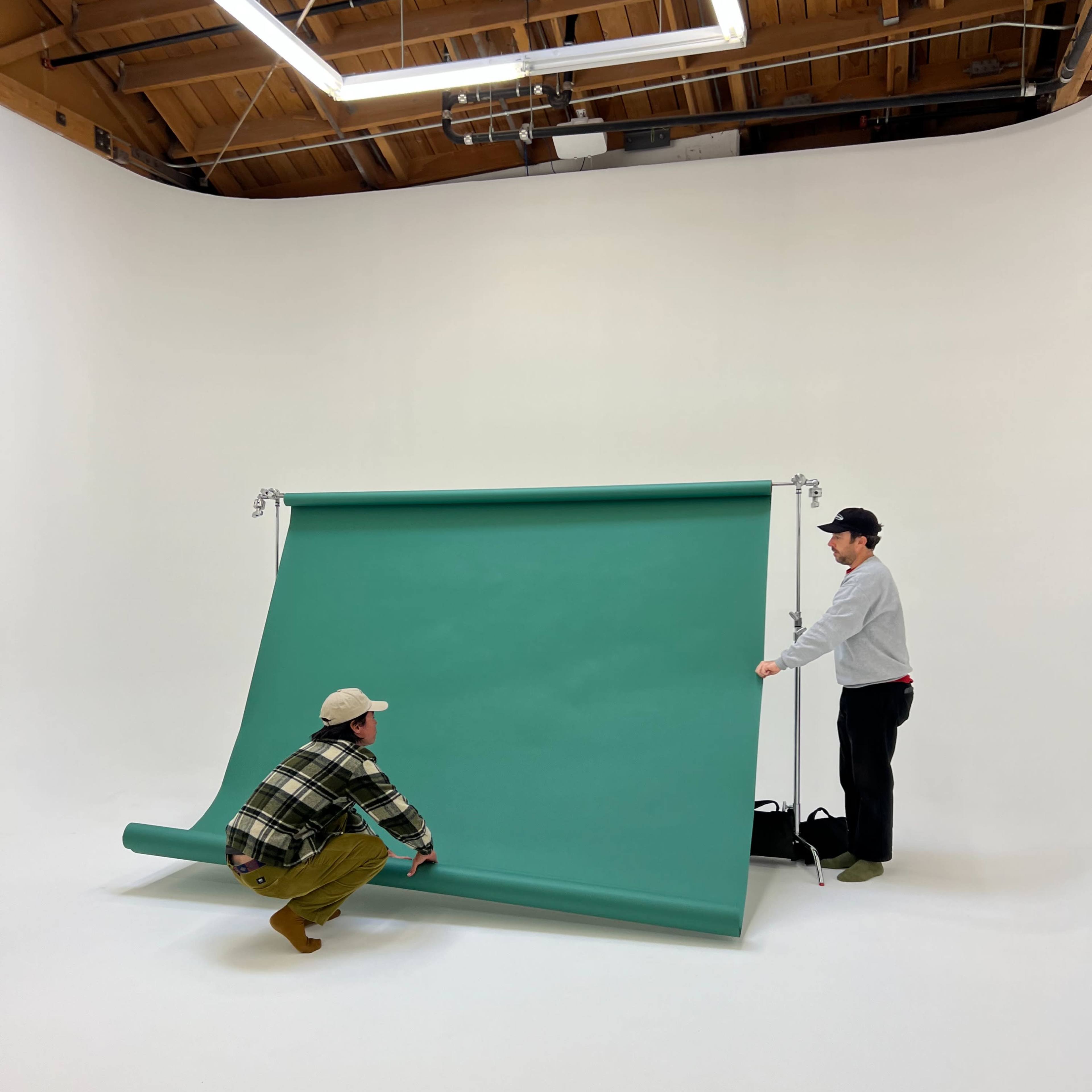 Two people are adjusting a green backdrop in a photography studio with a white background.