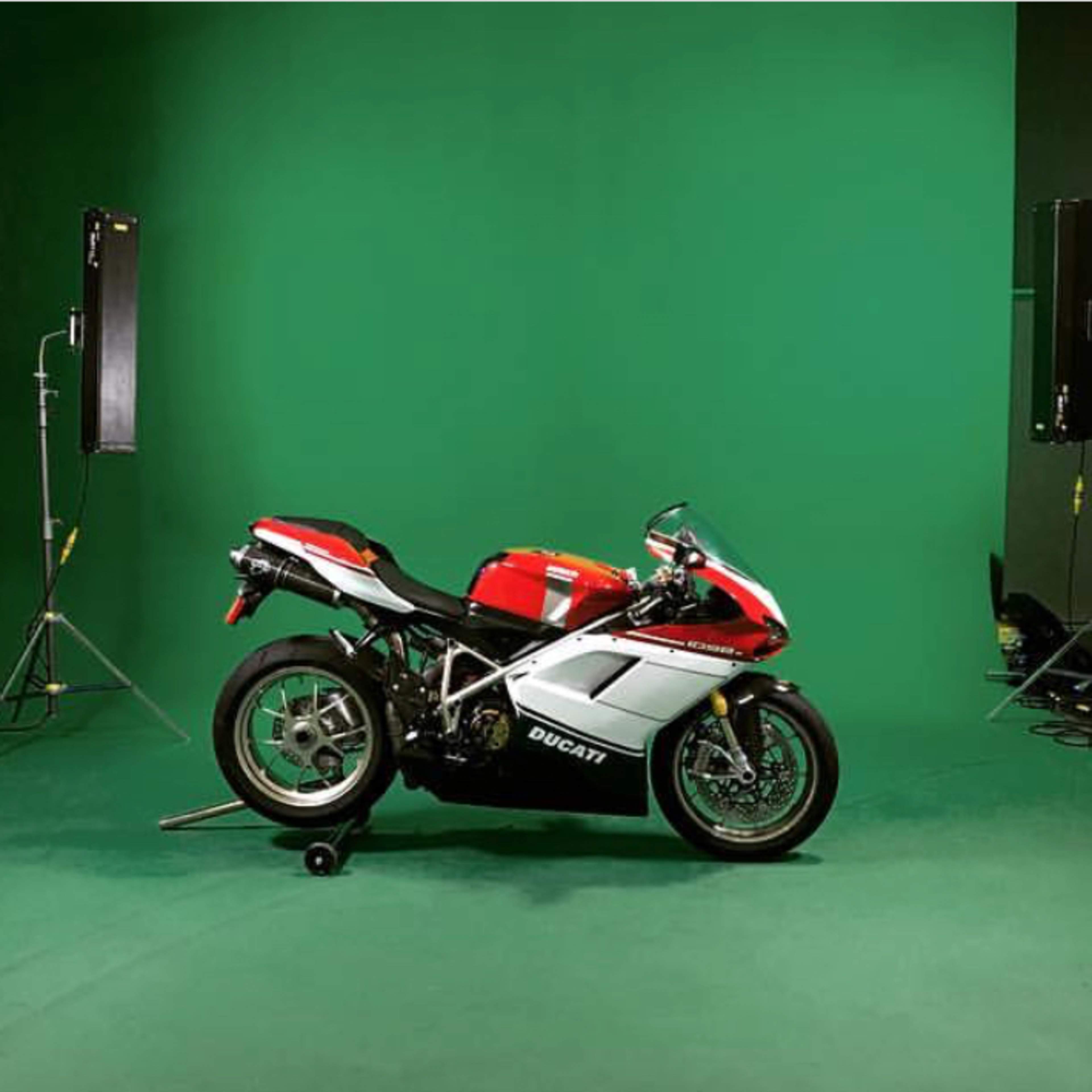 A Ducati motorcycle is positioned on a green backdrop in a studio, surrounded by lighting equipment and a small group of people.