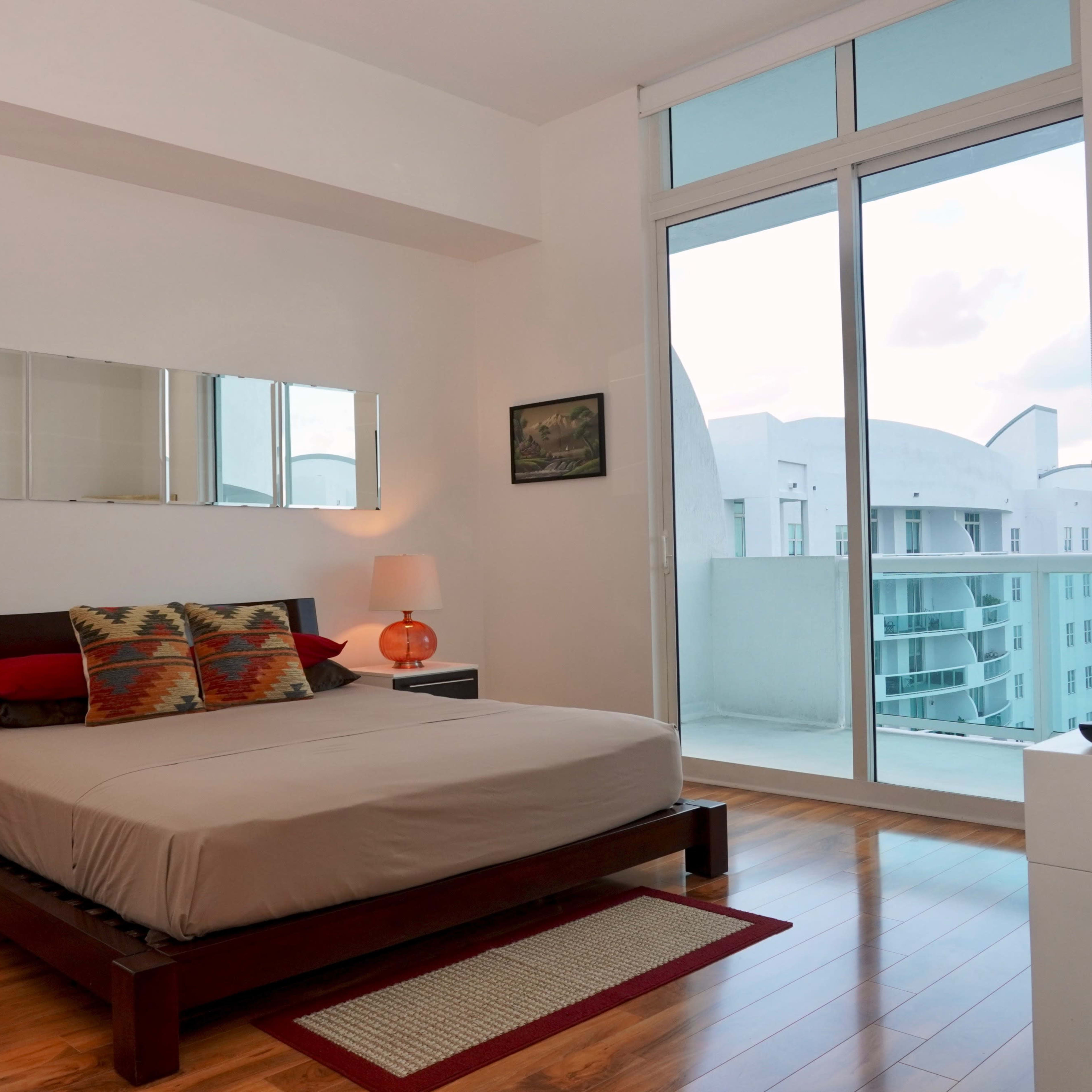 A modern bedroom features a bed with decorative pillows, two nightstands with lamps, and a large window overlooking a balcony.