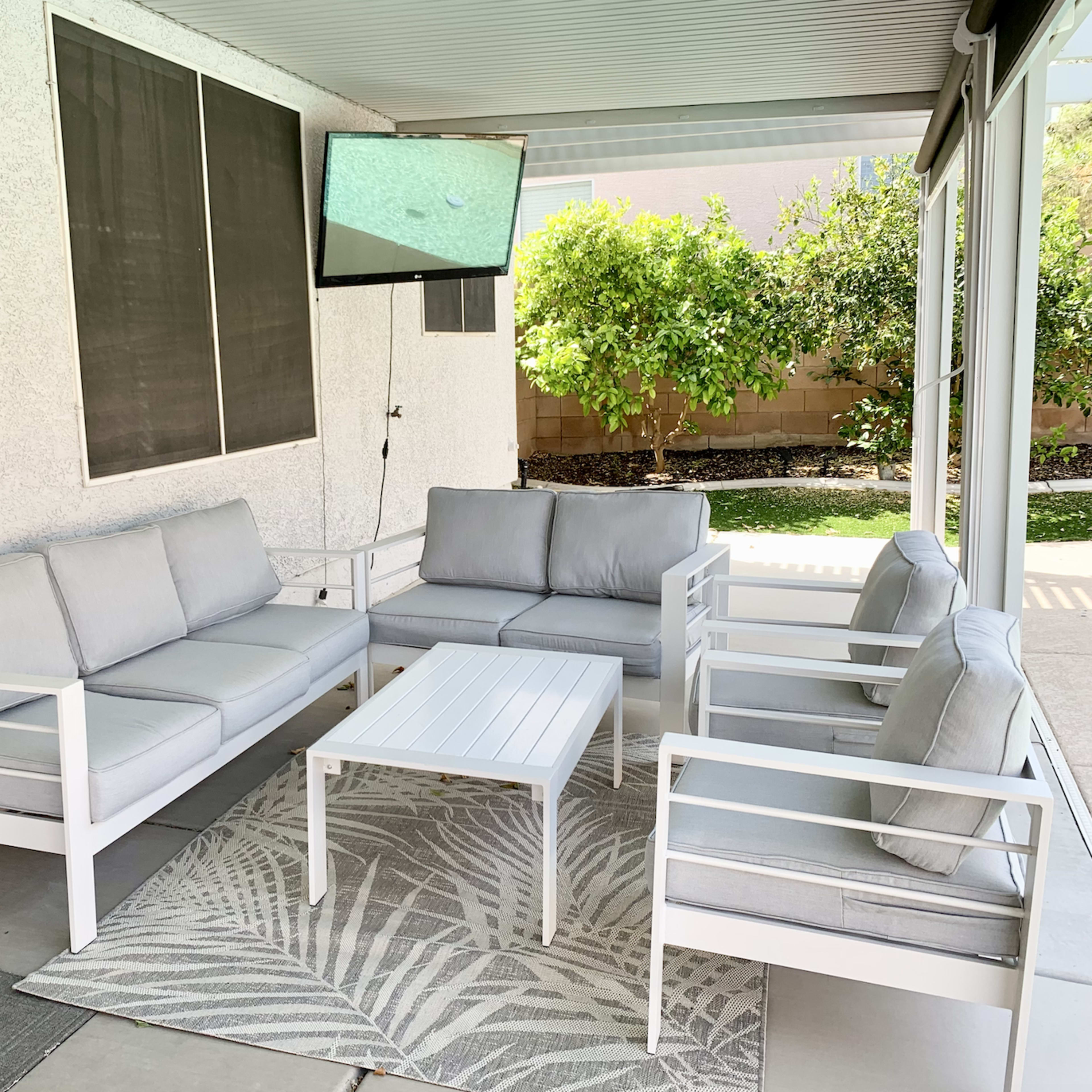 A patio area features white outdoor furniture arranged around a small coffee table, with a light rug underneath and greenery visible in the background.