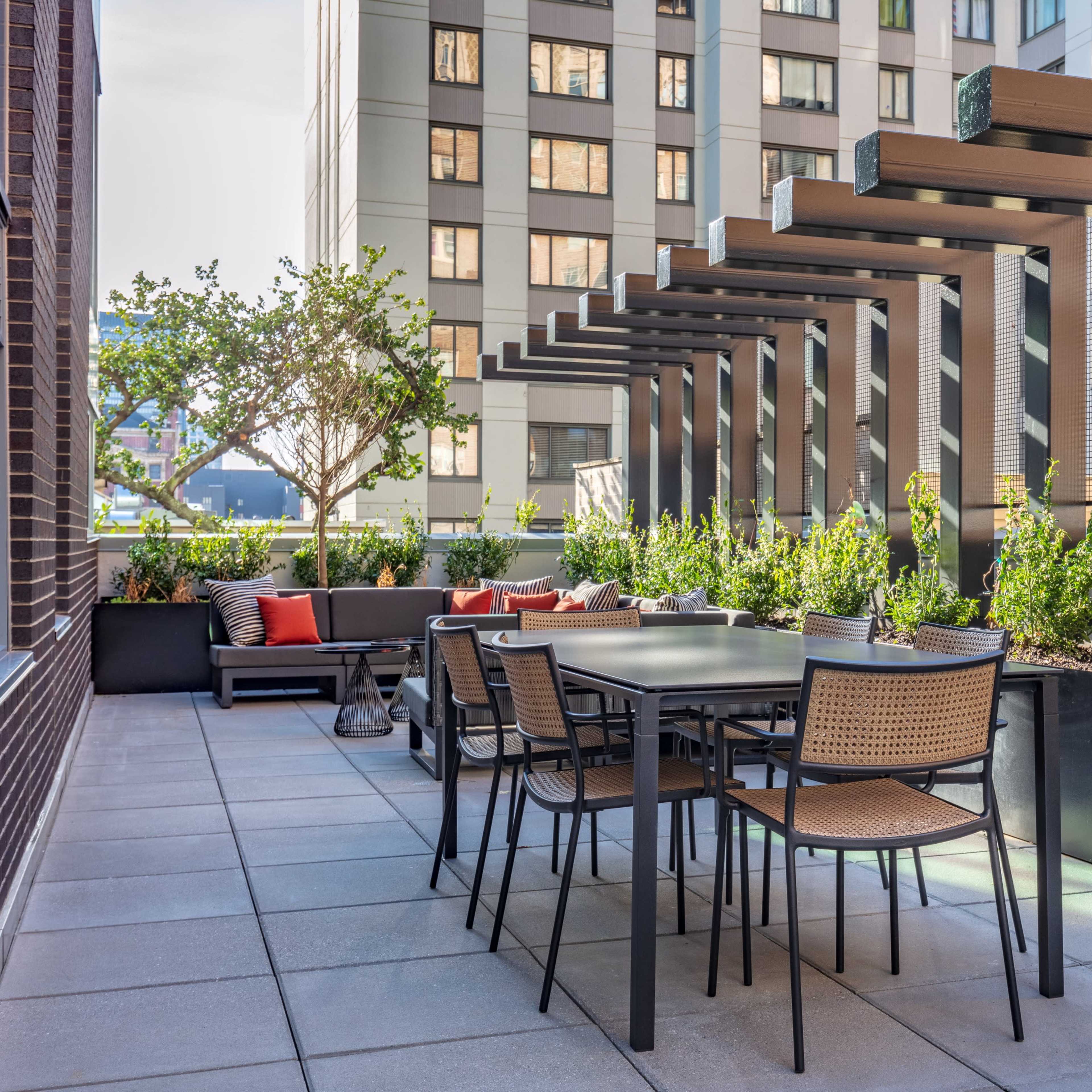 A patio area features a table and chairs surrounded by greenery and modern buildings.
