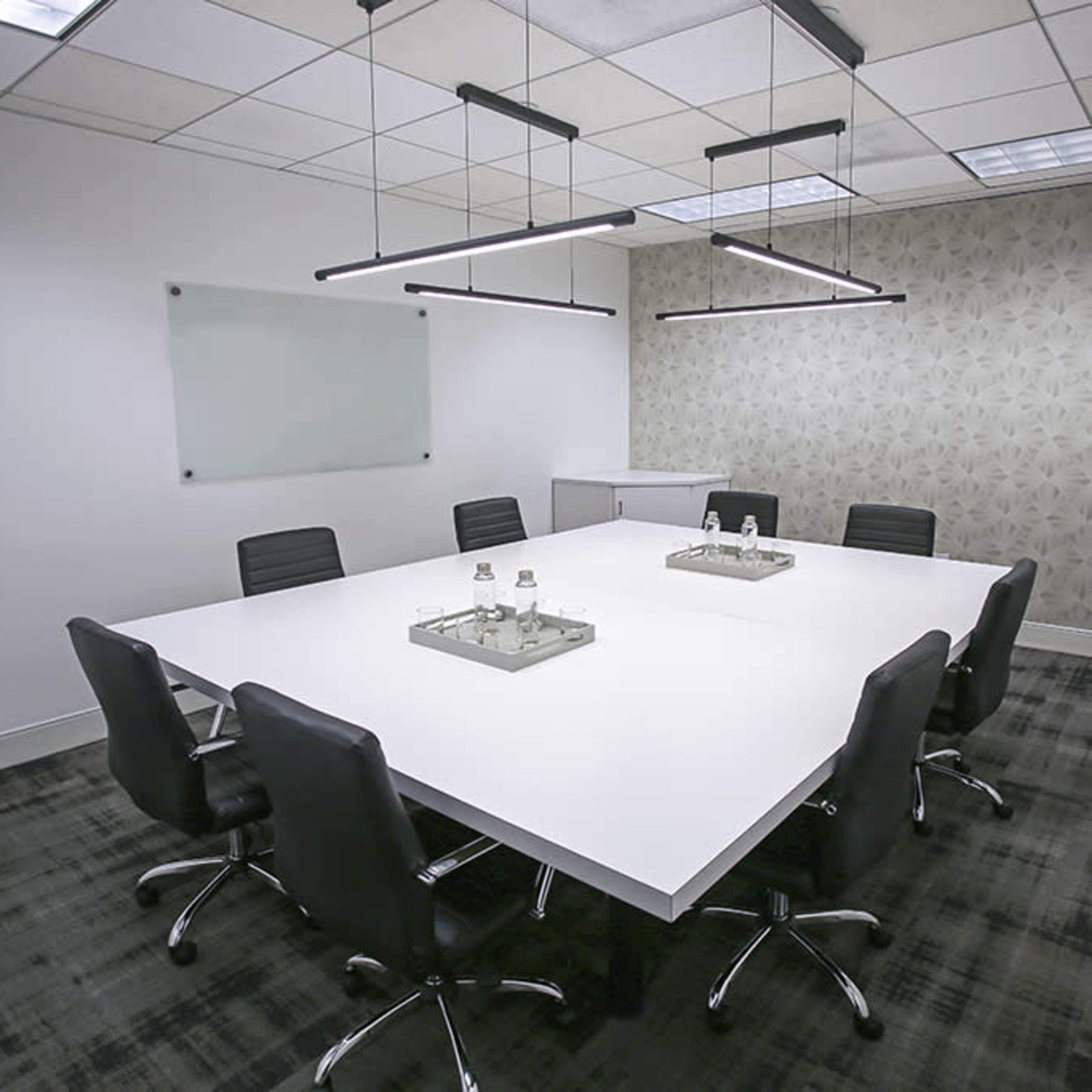 A modern conference room features a large white table surrounded by black chairs, with minimalistic light fixtures above and a patterned wall in the background.
