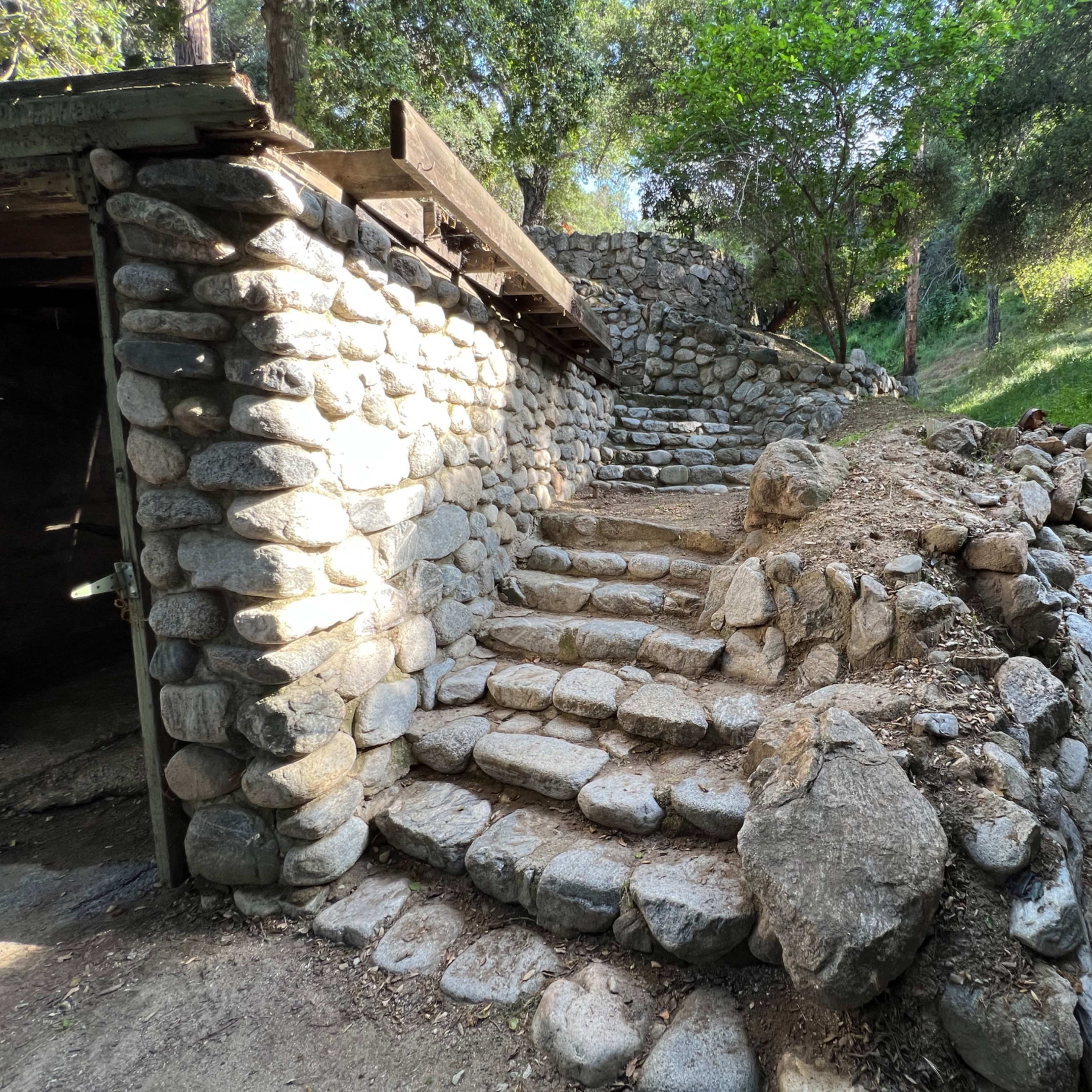 A stone pathway with steps leading up through a wooded area, flanked by stone walls and surrounding greenery.