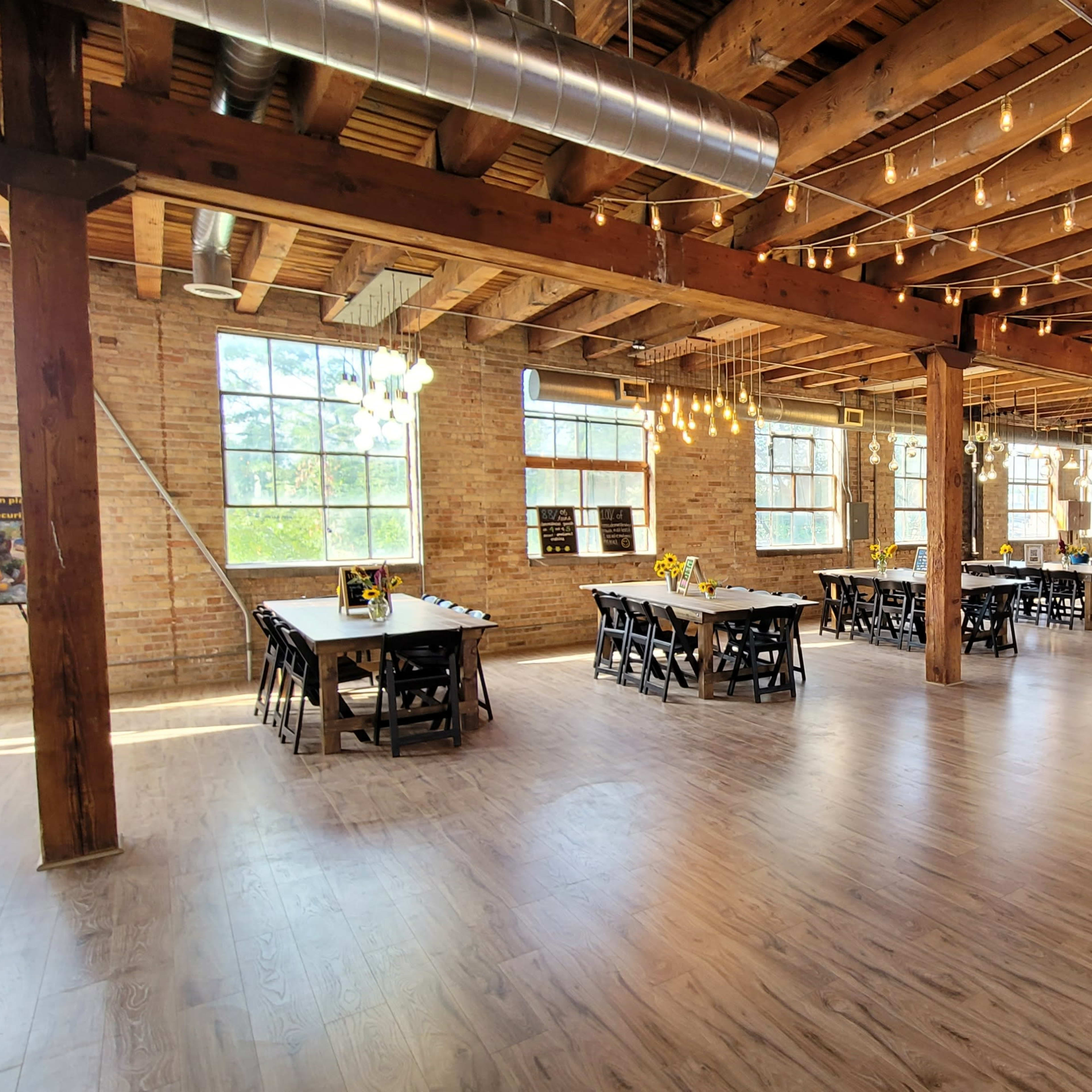 A spacious, well-lit event venue with wooden beams, industrial-style lighting, and rows of tables set up for a gathering.