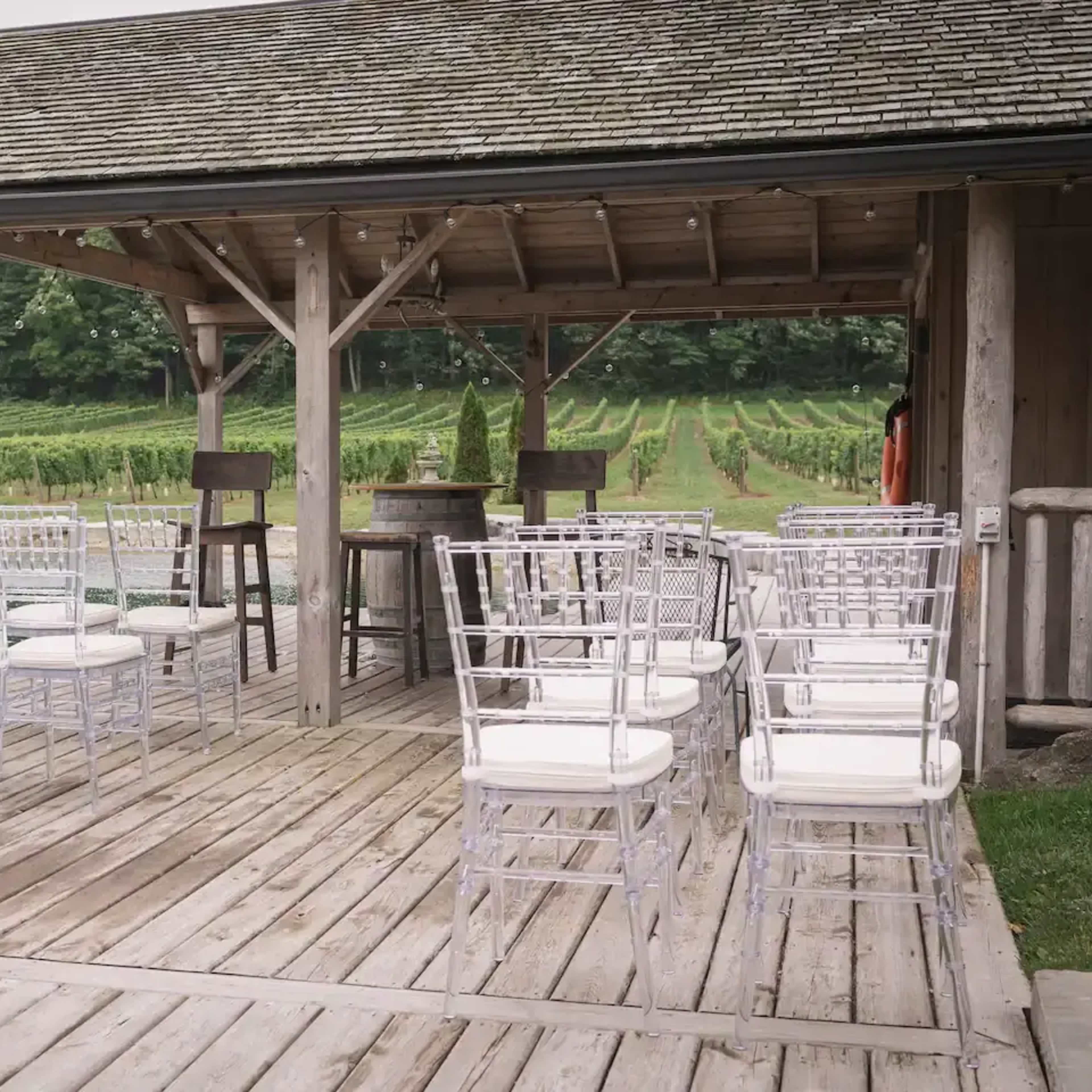 A wooden pavilion set up for an outdoor event, featuring rows of clear chairs with white cushions overlooking a vineyard.