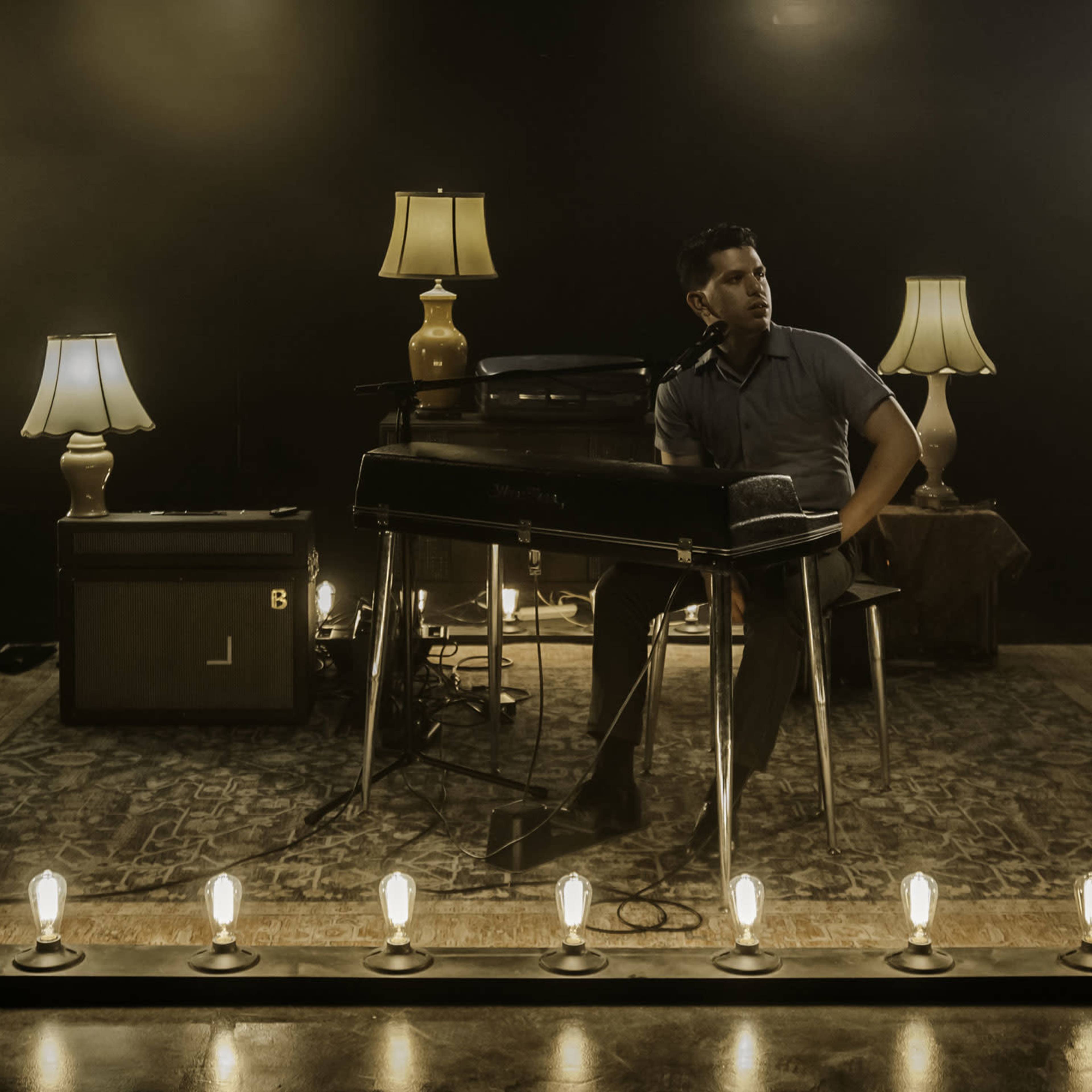 A musician sits at a keyboard on a rug surrounded by several lamps and illuminated bulbs in a dimly lit room.