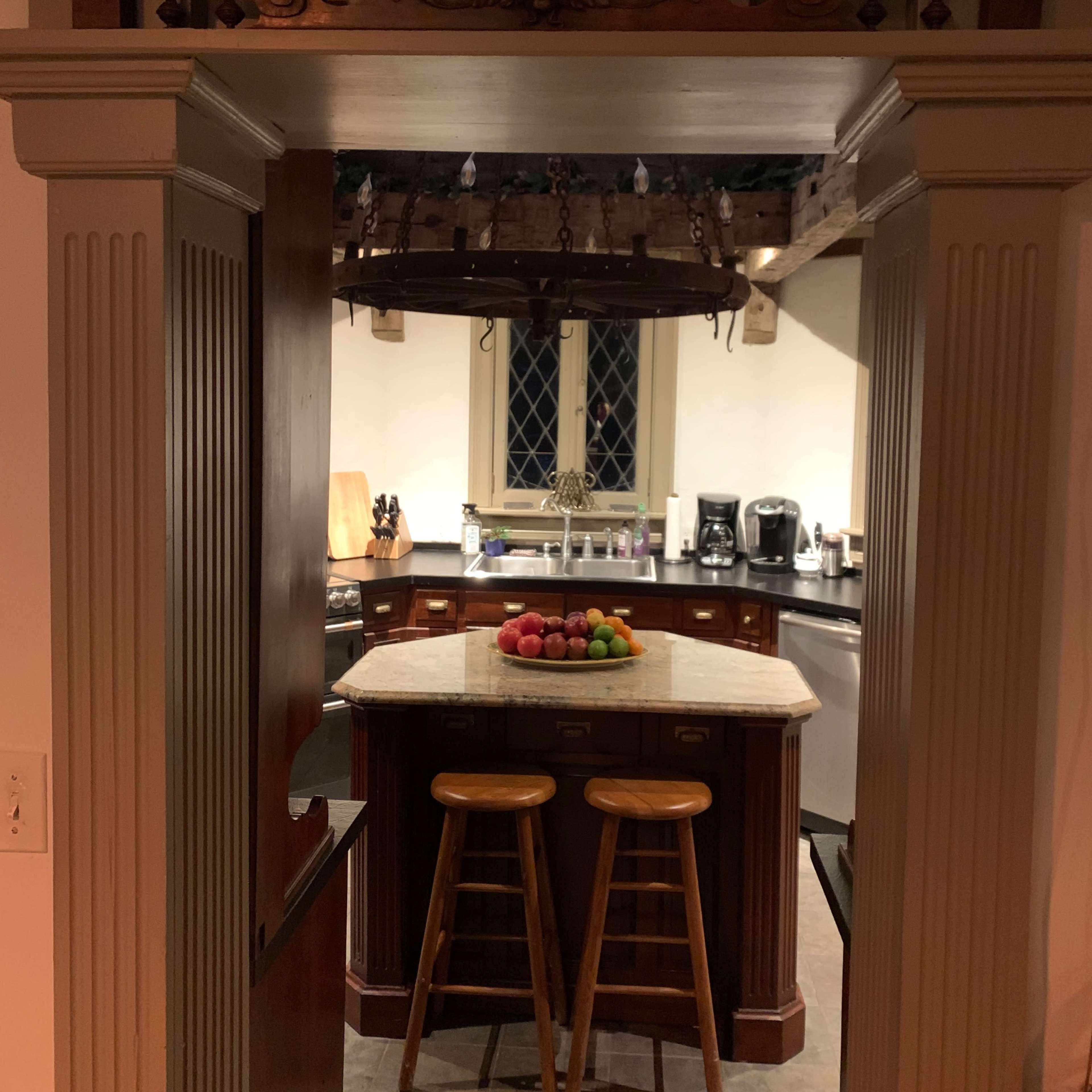 A view through an archway into a kitchen featuring a central island with two wooden stools and a bowl of fruit on it.