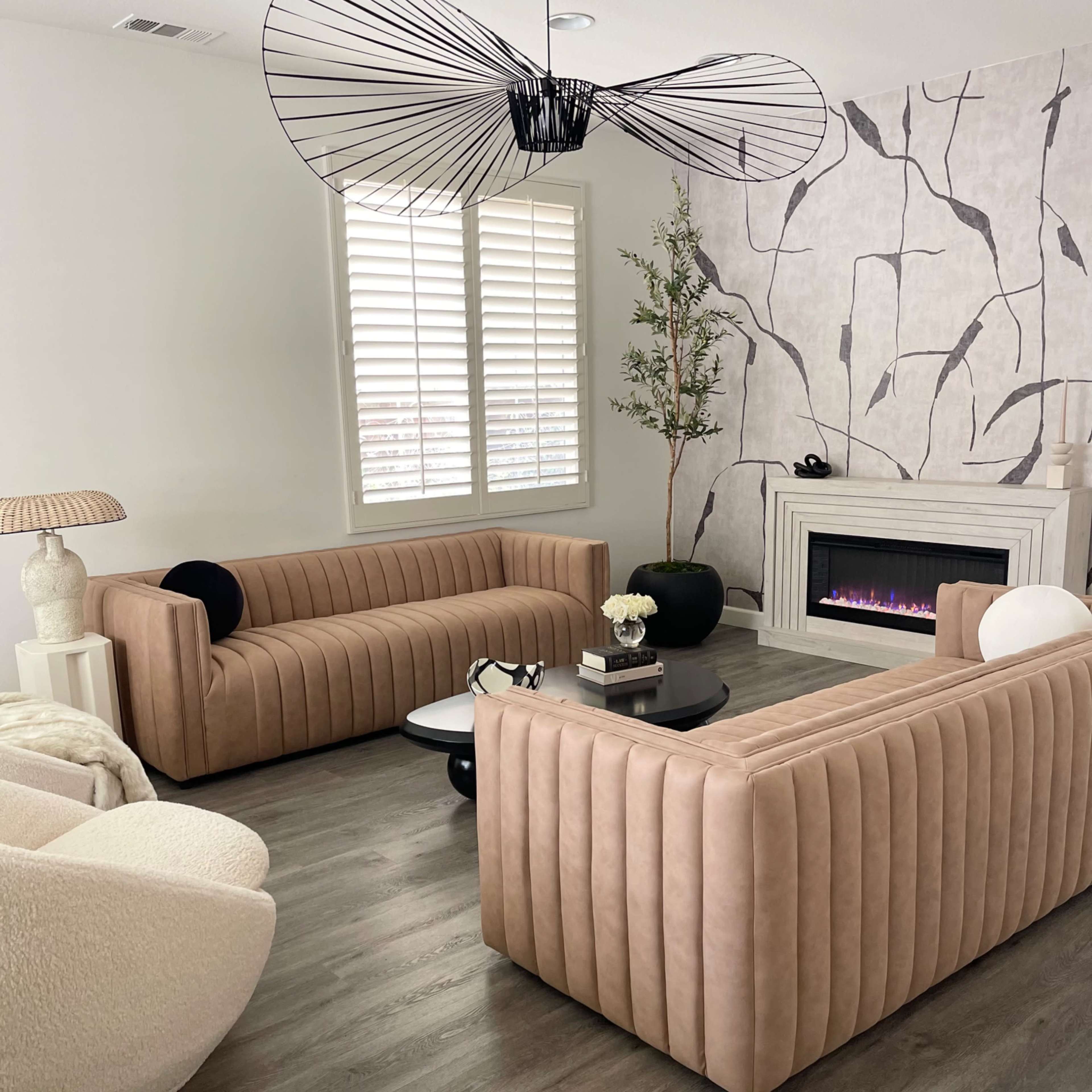 A modern living room features two tufted beige sofas facing each other, accompanied by a round black coffee table and a cozy armchair, with a decorative wall and a gas fireplace in the background.