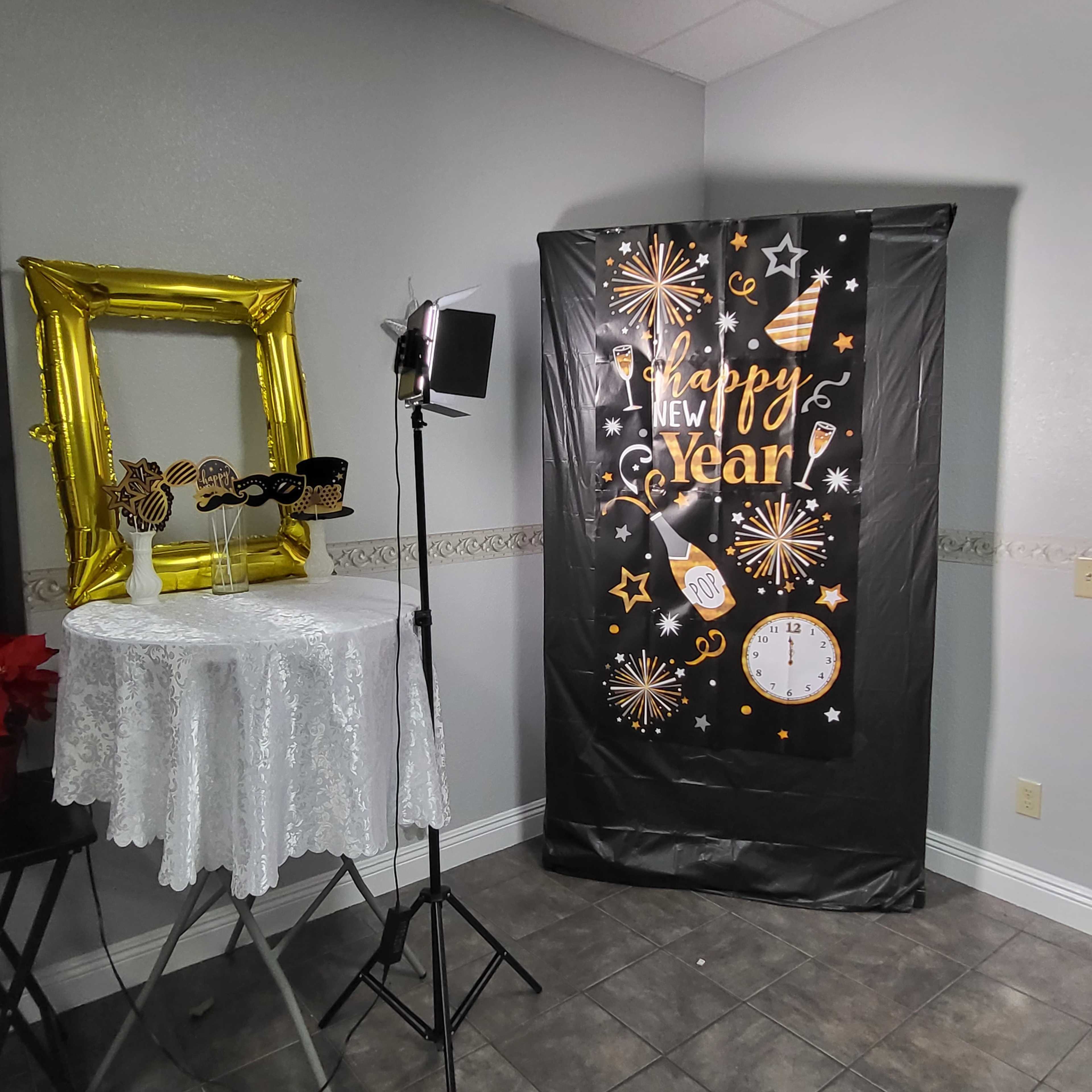 A decorated corner of a room with a "Happy New Year" backdrop, a table with a lace tablecloth, and a mirrored frame alongside party glasses.