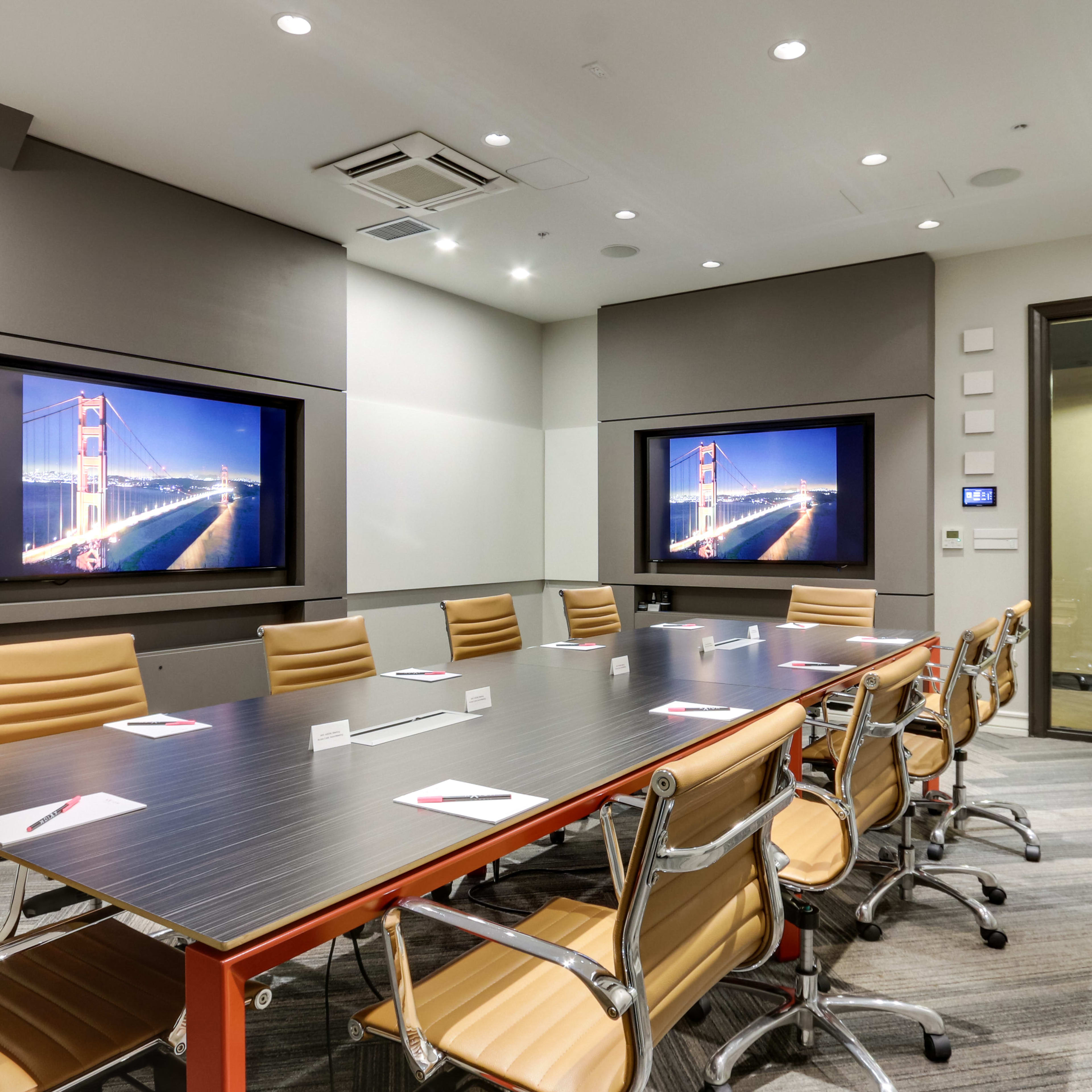 A modern conference room features a long table surrounded by ergonomic chairs, with two large screens displaying an illuminated bridge at night.