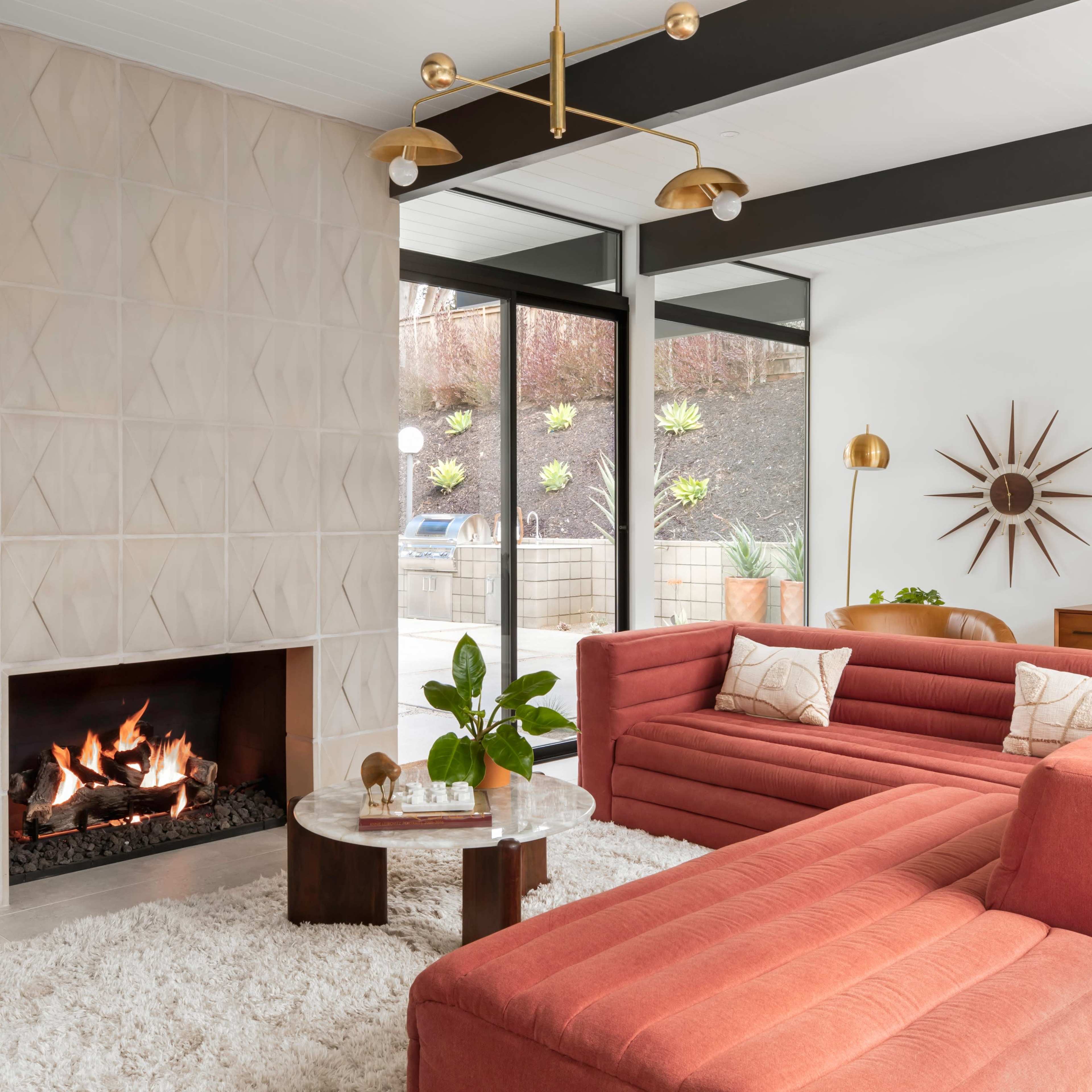 A mid-century modern living room features a coral sectional sofa, a circular coffee table, a fireplace, and a workspace with a computer, all surrounded by large windows.