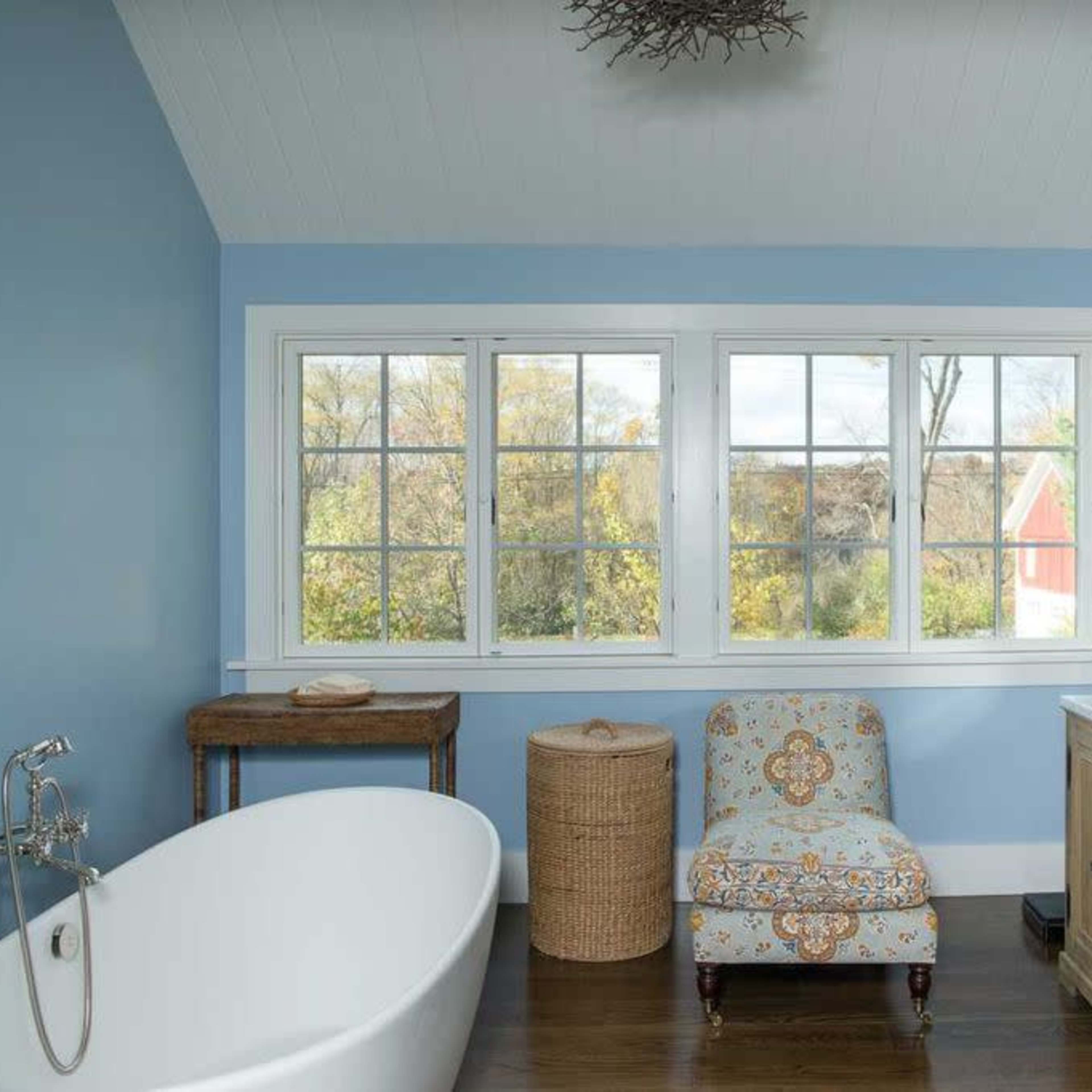 A bathroom features a freestanding bathtub, a pair of sinks, a decorative chair, and large windows with a view of trees.