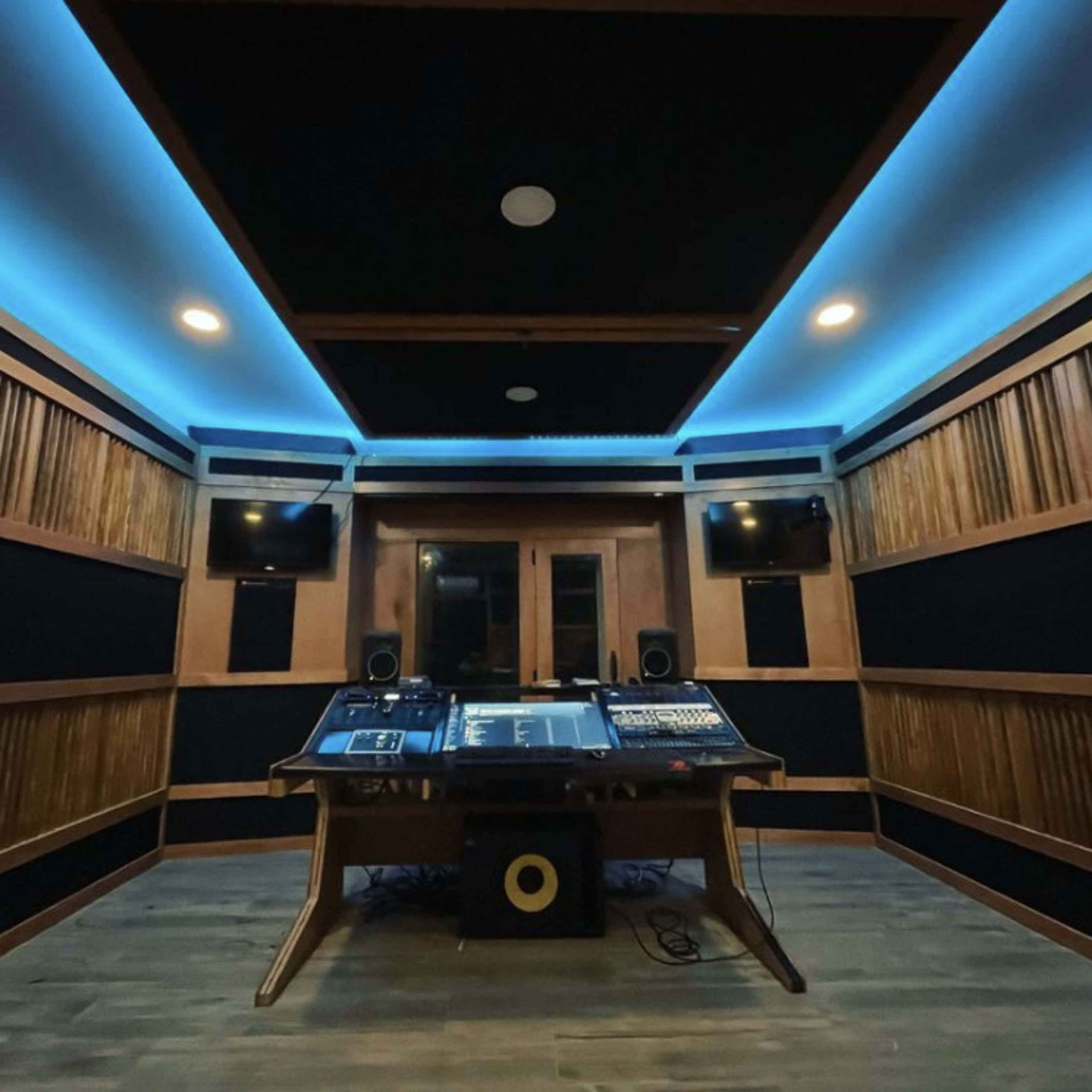 A modern recording studio with wooden walls, a sound mixing desk in the center, and blue LED lights illuminating the ceiling.
