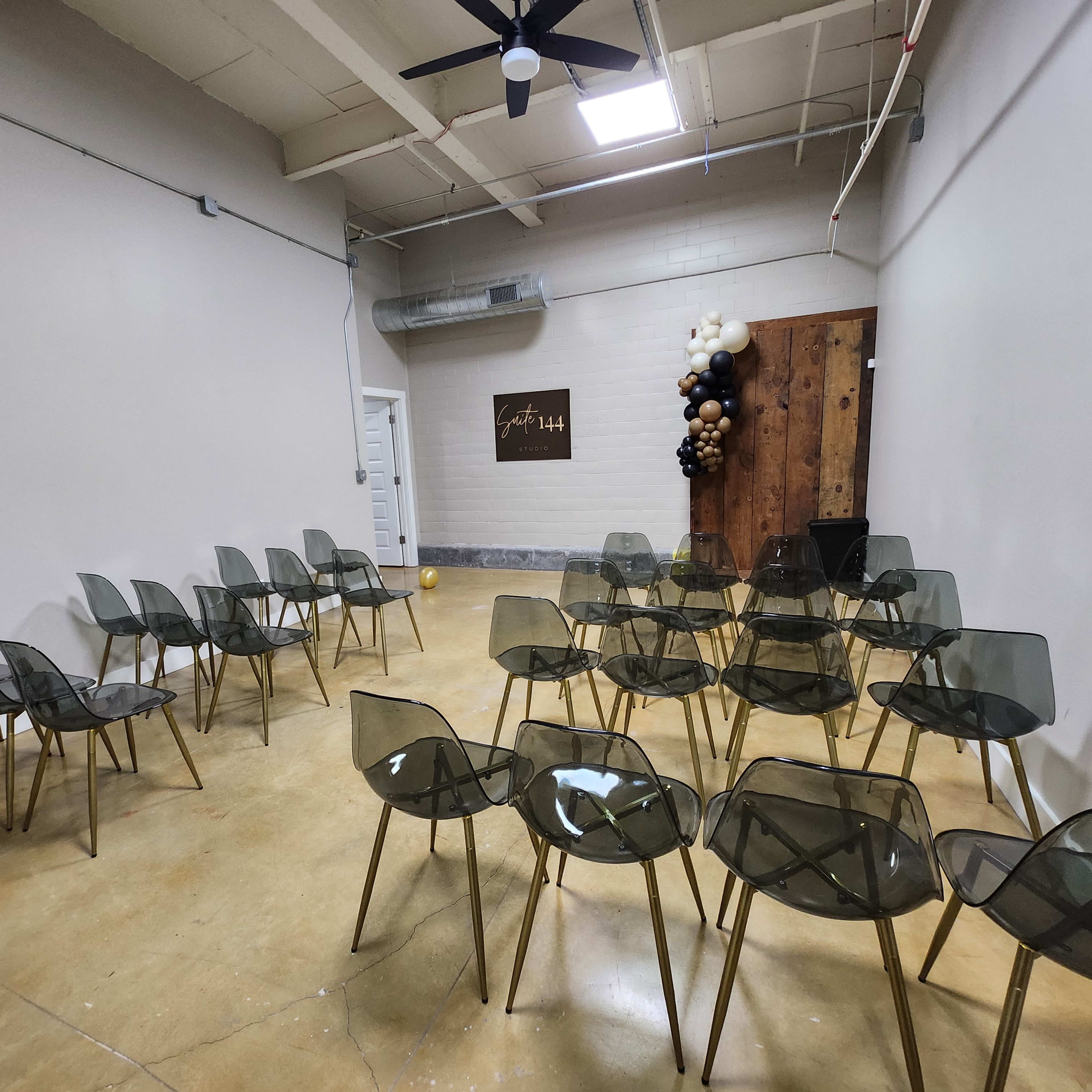 A minimalist event space with transparent chairs arranged in rows facing a wall decorated with a wood panel and a balloon arrangement.