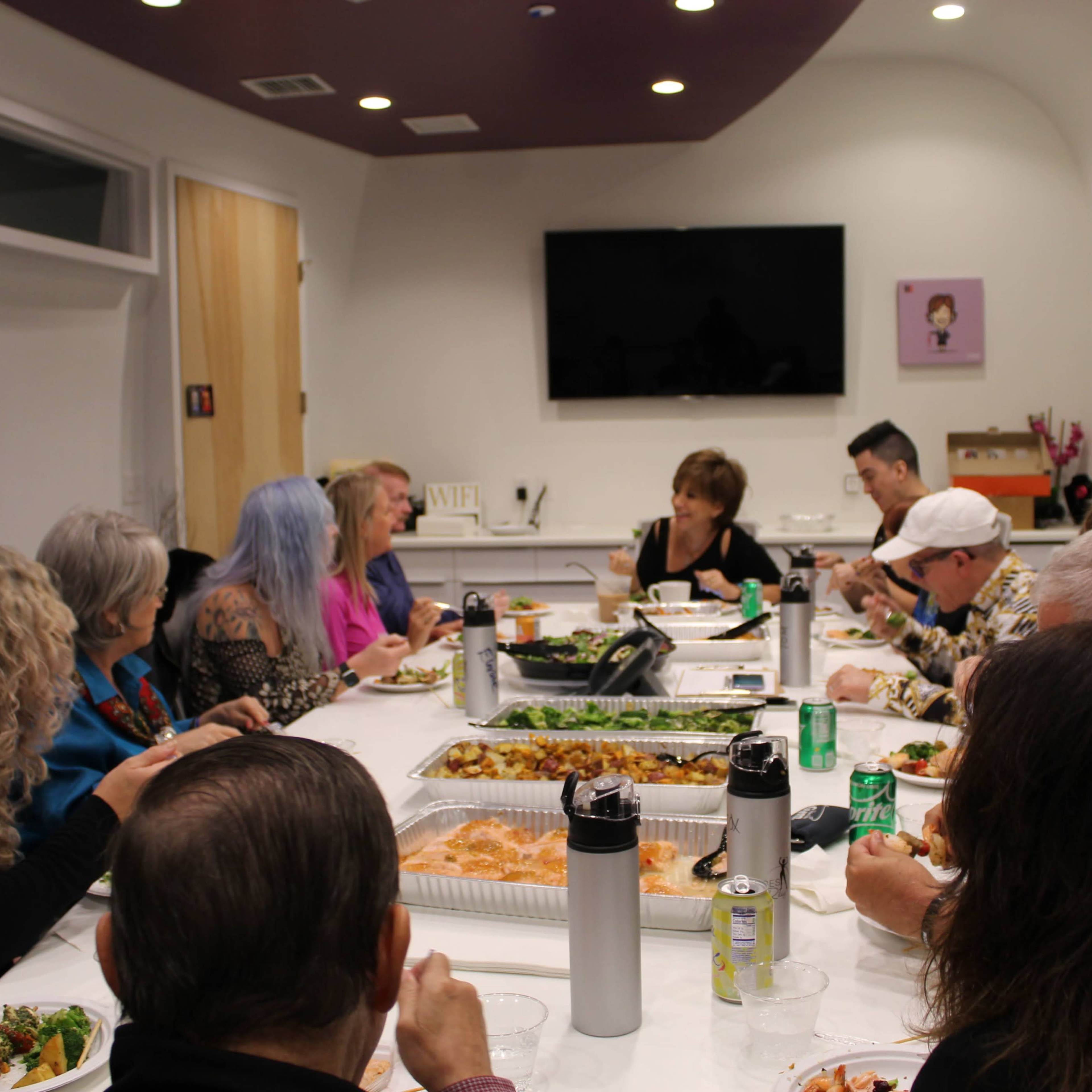 A group of people is seated around a long table, enjoying a meal together in a brightly lit meeting room.