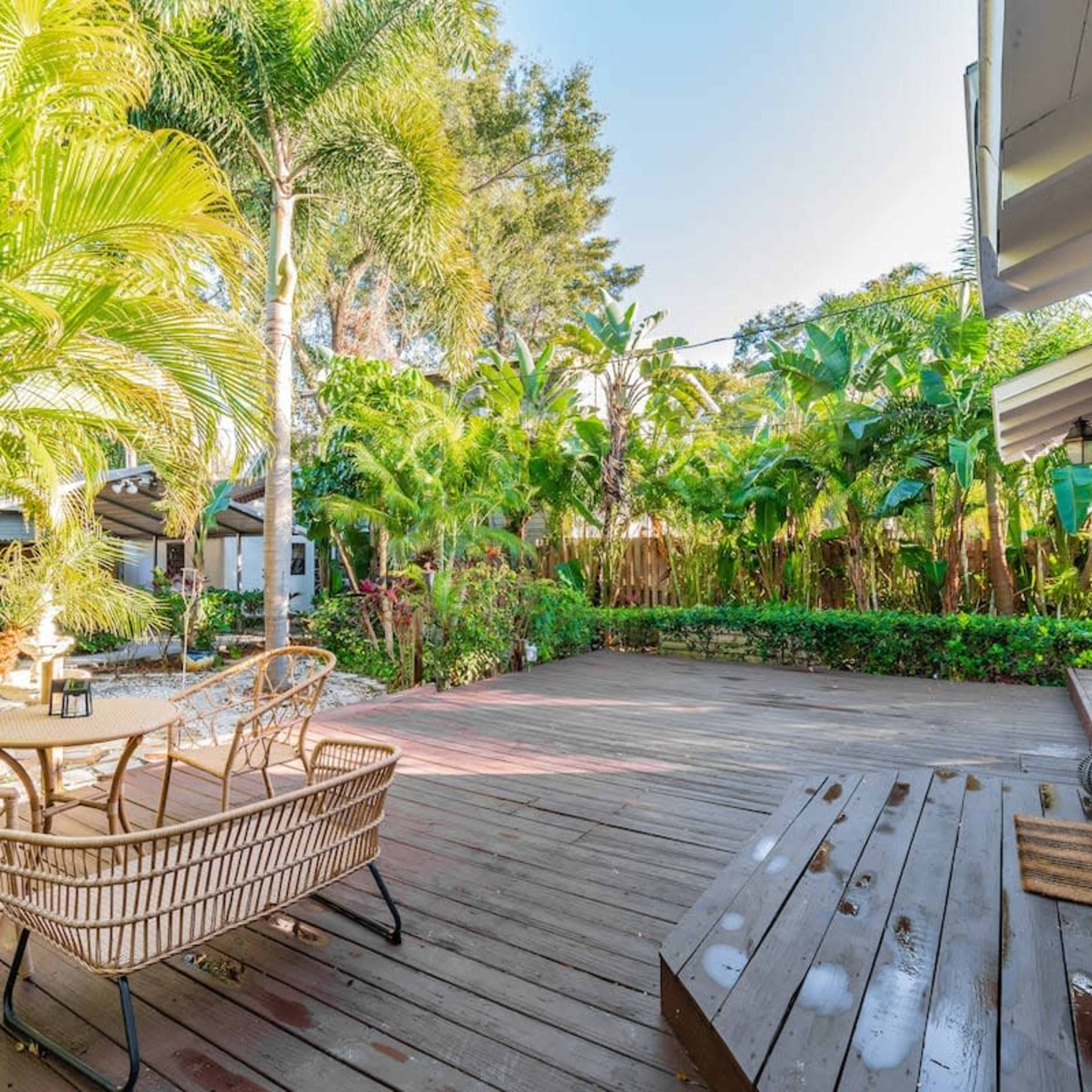 A wooden deck surrounded by lush greenery and tropical plants, furnished with a table and chairs.