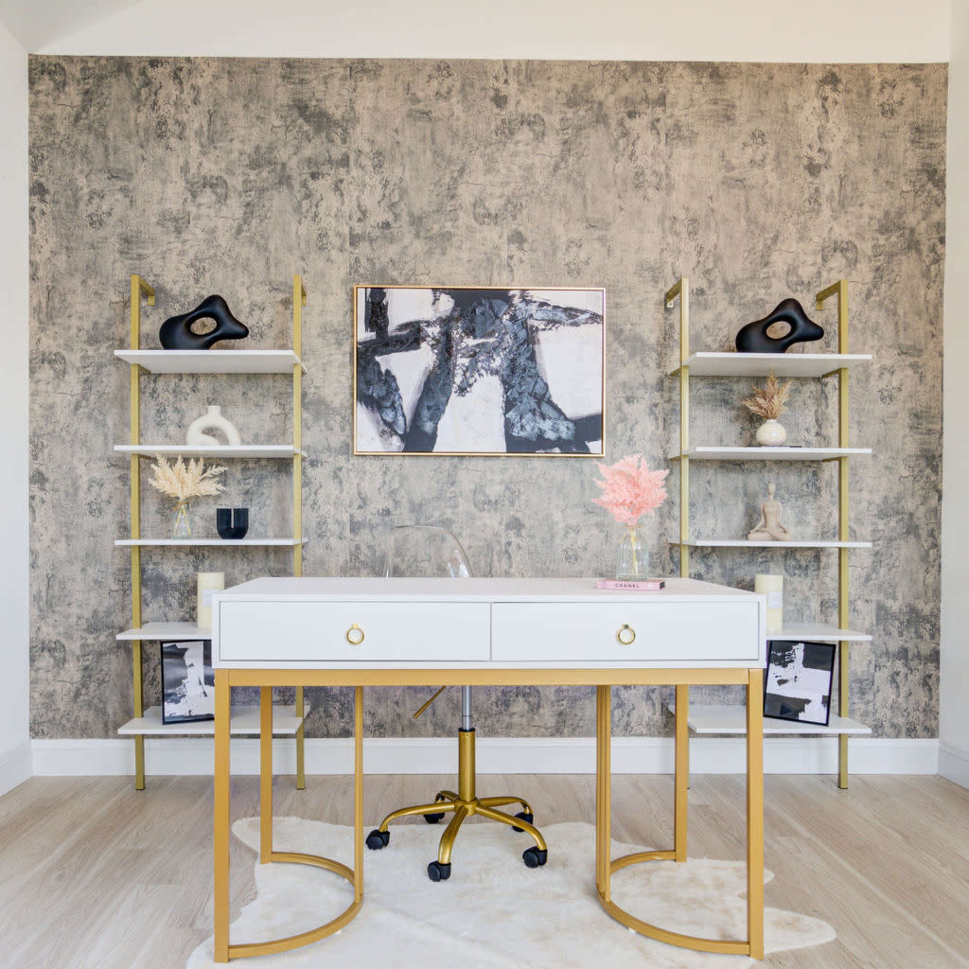 A modern office space features a white desk with gold accents, surrounded by two vertical shelves holding decorative items, against a textured gray wall.