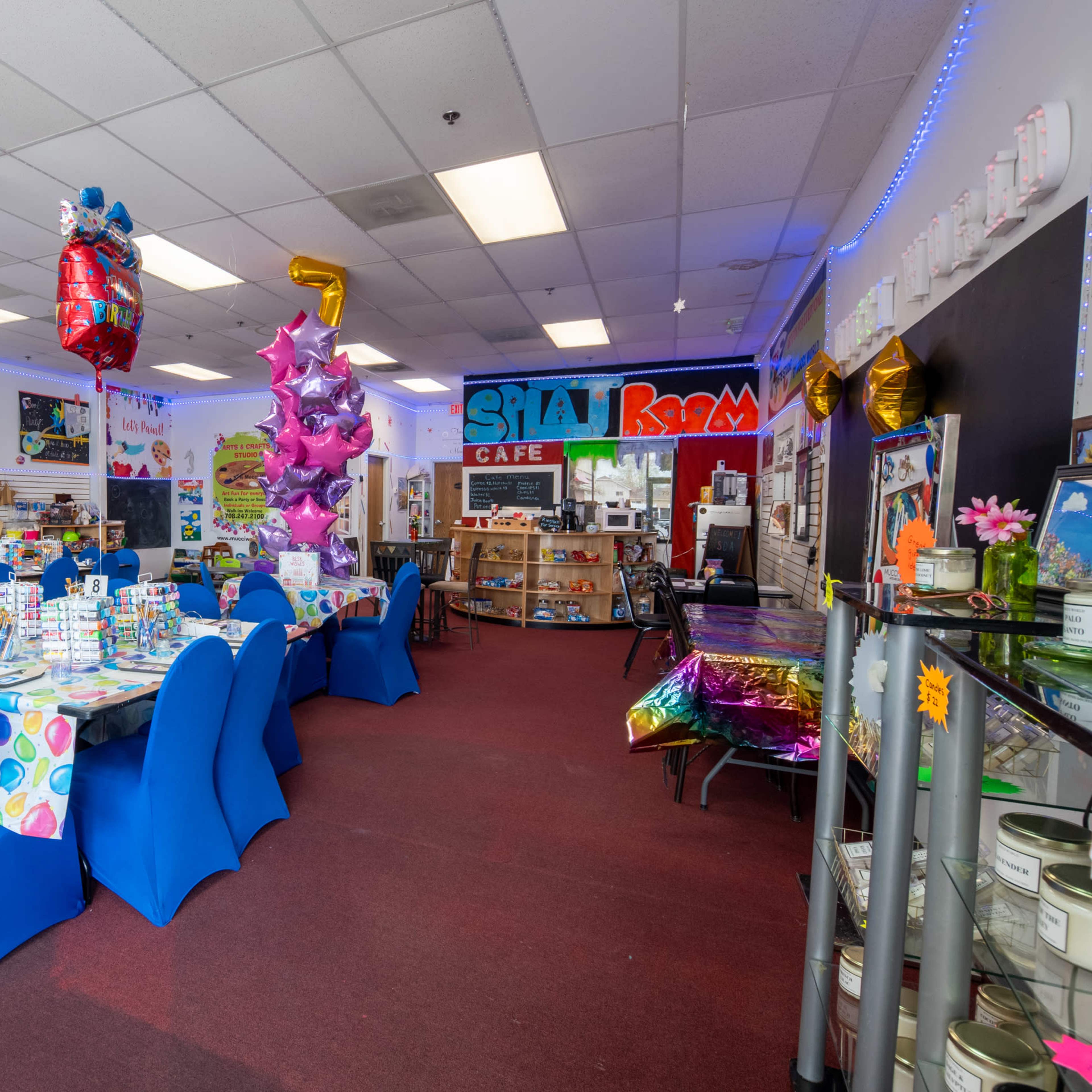 A colorful café interior with blue chairs and tables covered in festive tablecloths, decorated with balloons and various party supplies.
