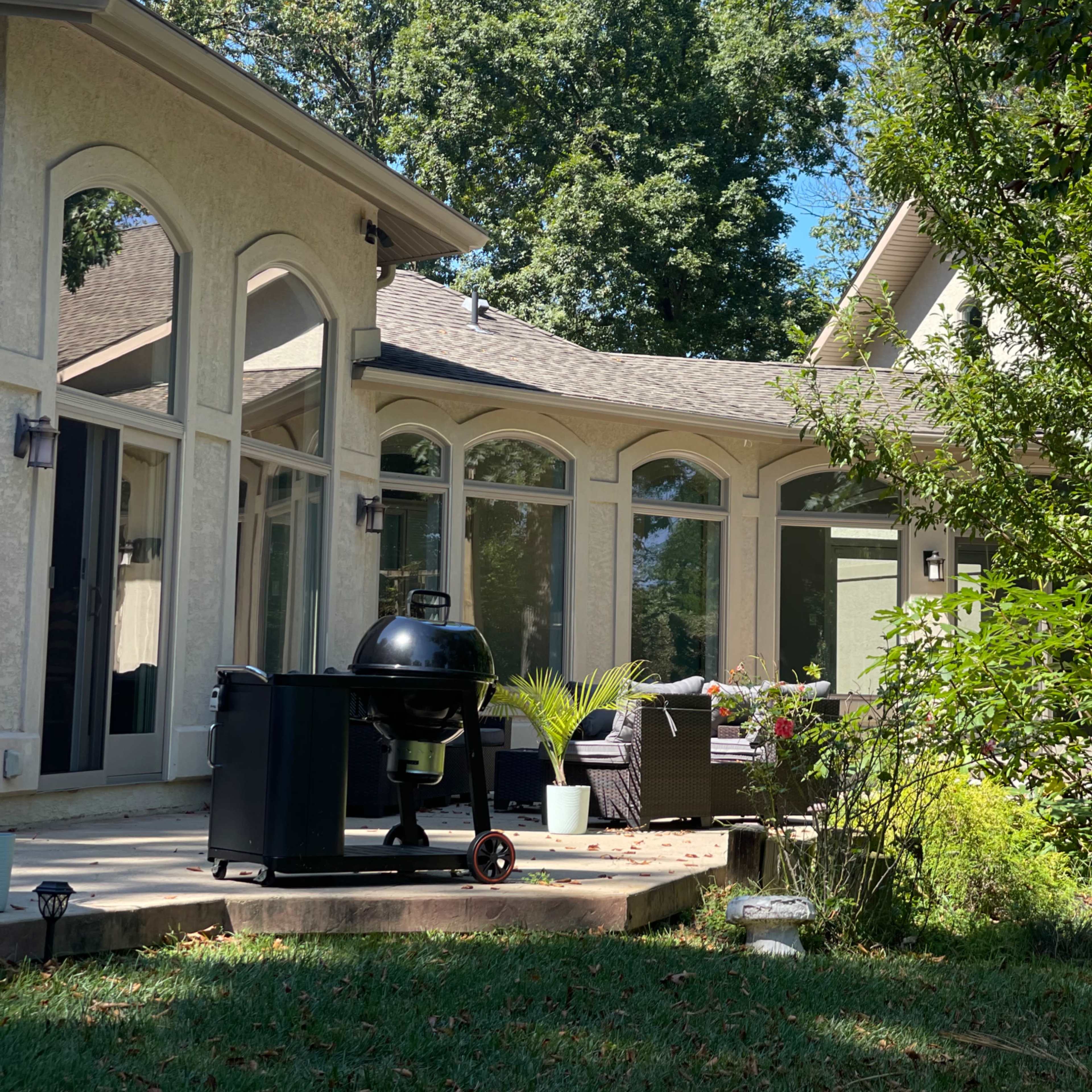 A patio with a charcoal grill and outdoor seating is situated next to a house surrounded by trees.