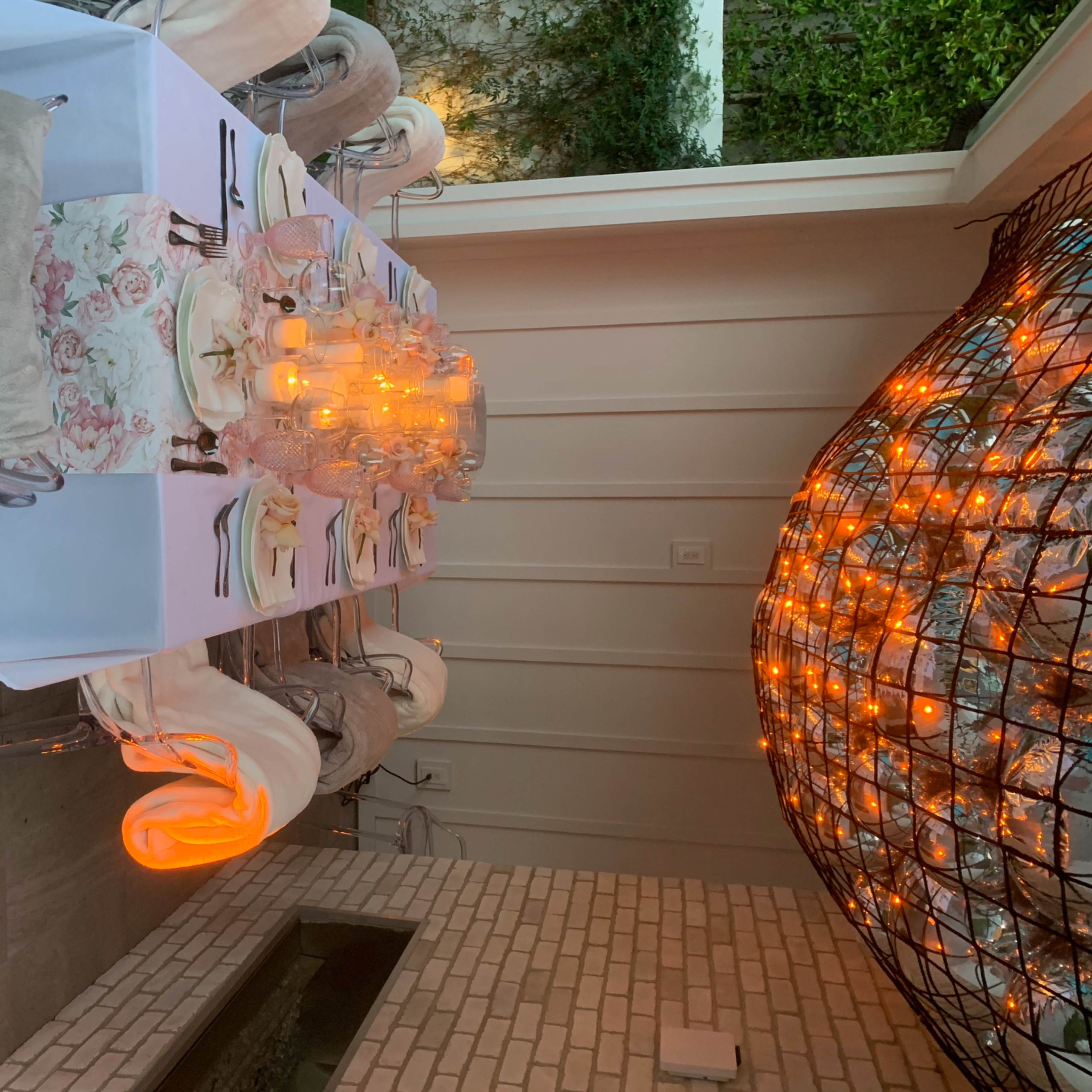 A long dining table is set with elegant place settings and glowing candles under a modern light fixture in a stylish indoor-outdoor space.