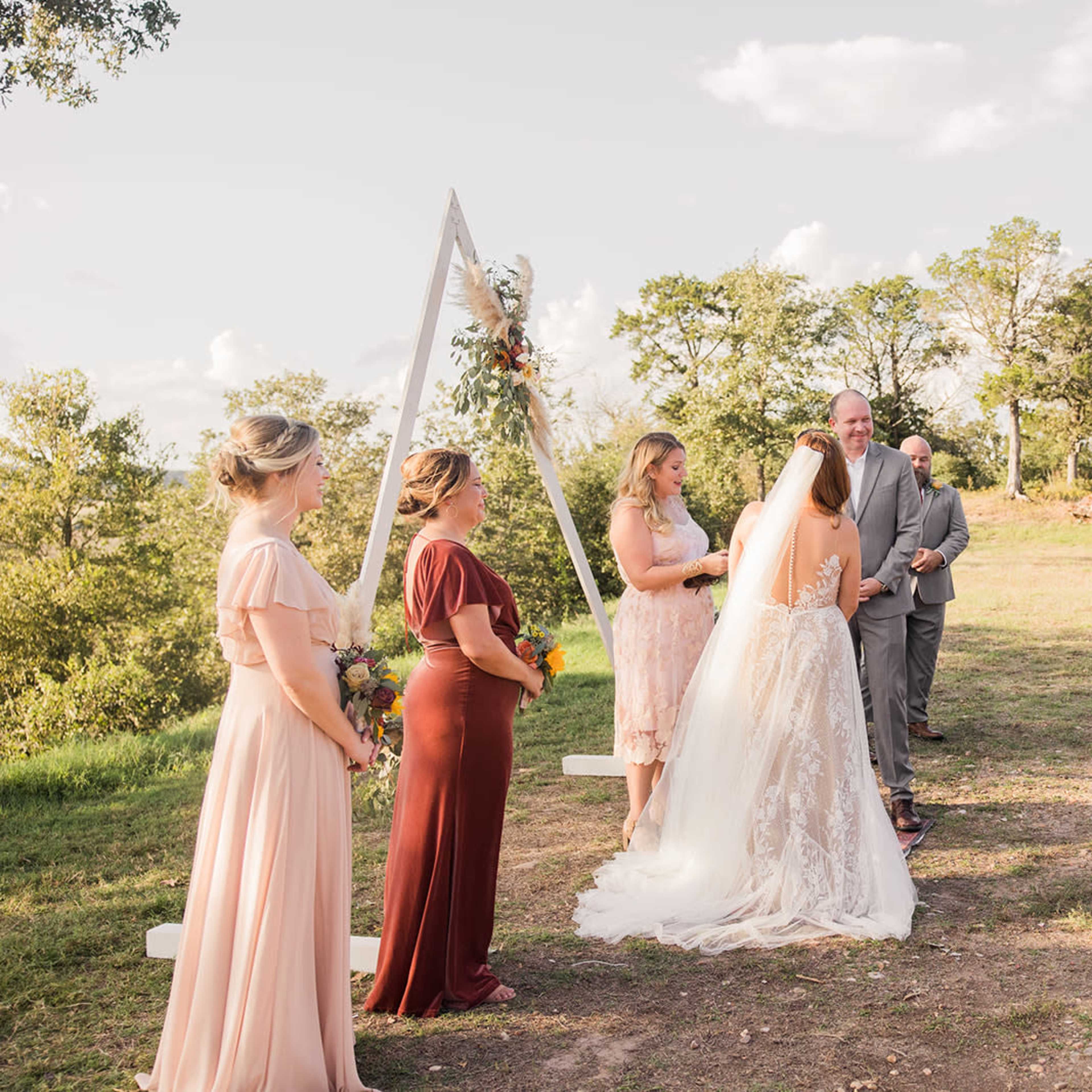 A couple exchanges vows during an outdoor wedding ceremony, surrounded by guests seated on either side and a decorated arch in the background.