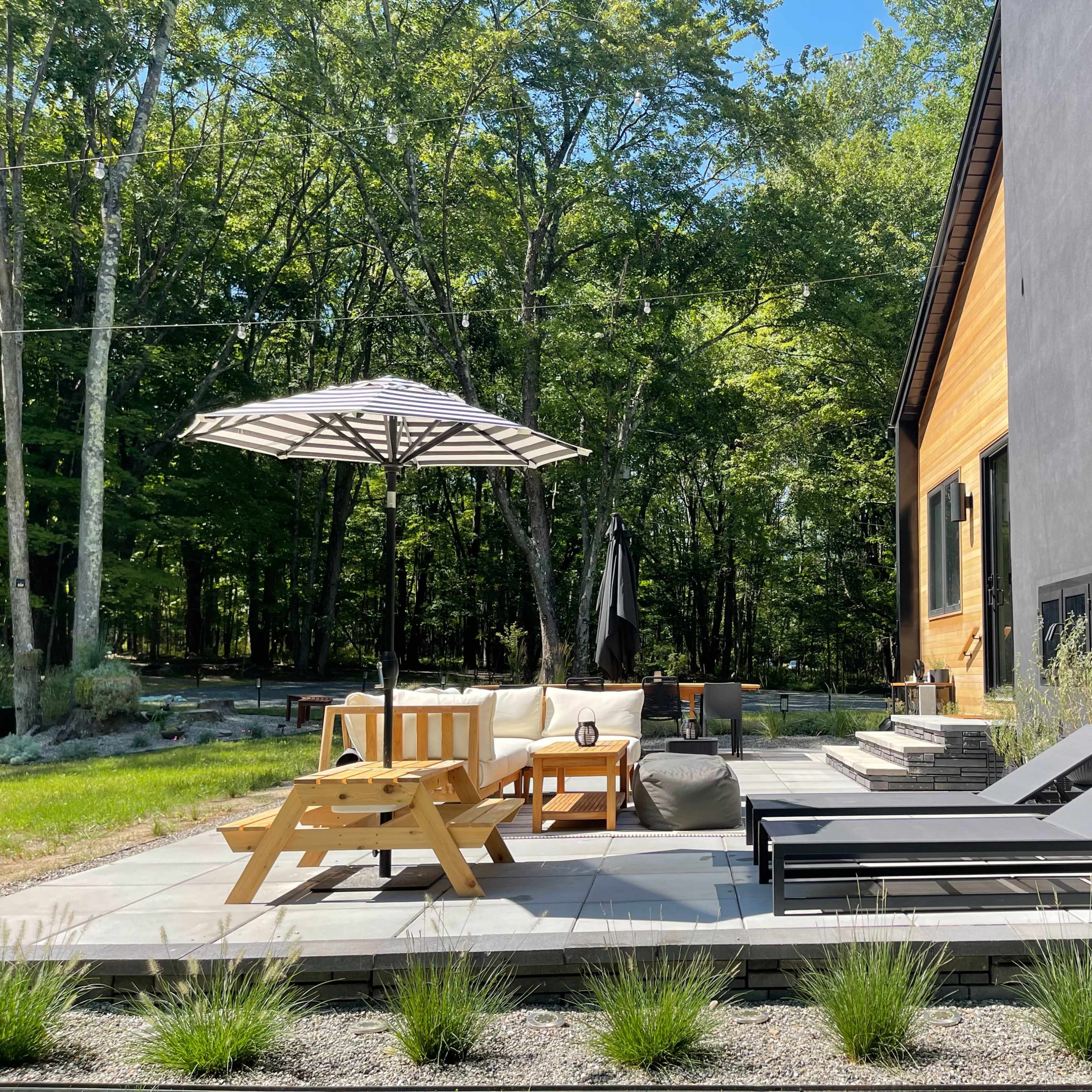 A patio area features a wooden lounge set, an umbrella, and two chaise loungers surrounded by trees.