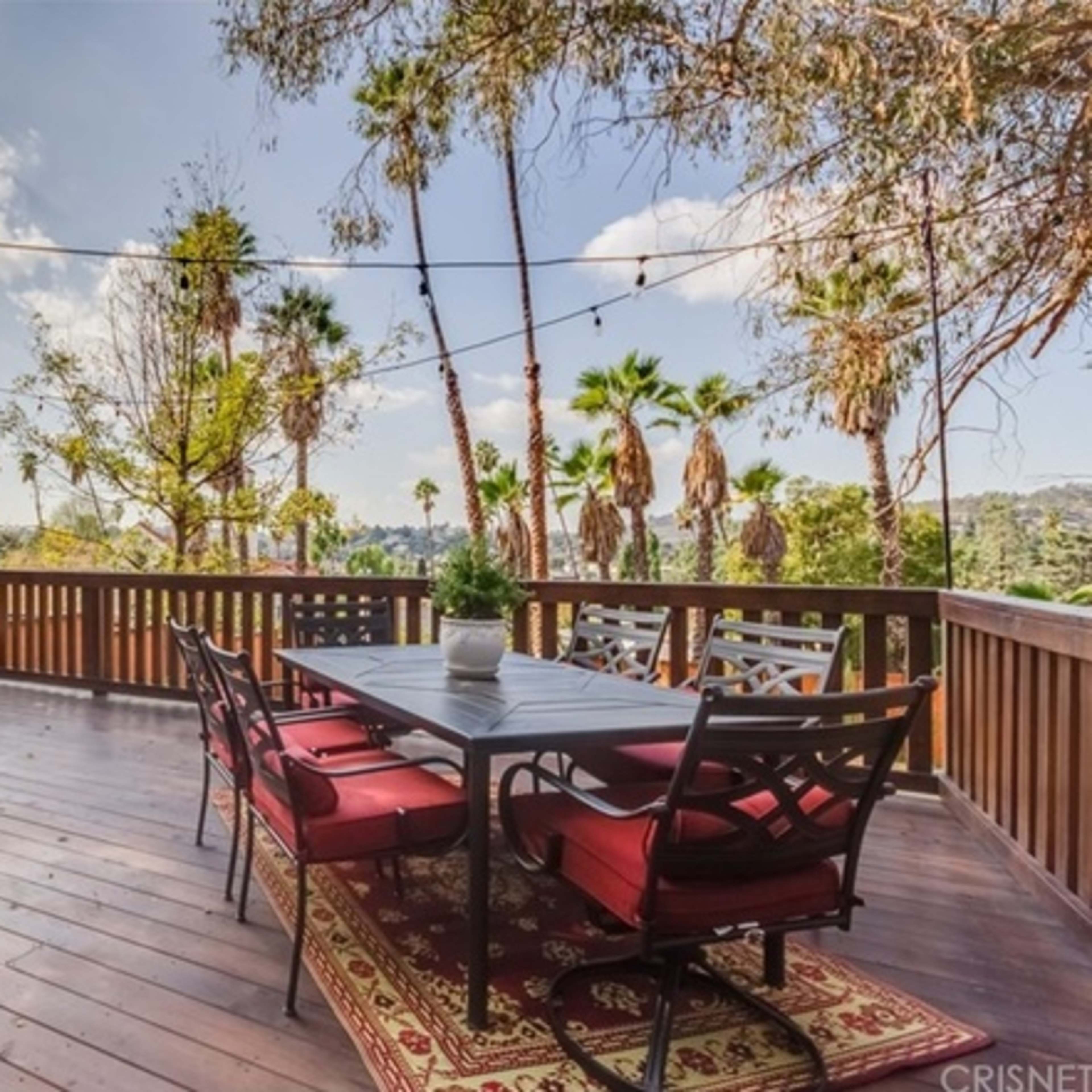 A wooden deck features a dining table with black chairs and a potted plant, surrounded by palm trees and a mountainous backdrop under a partly cloudy sky.