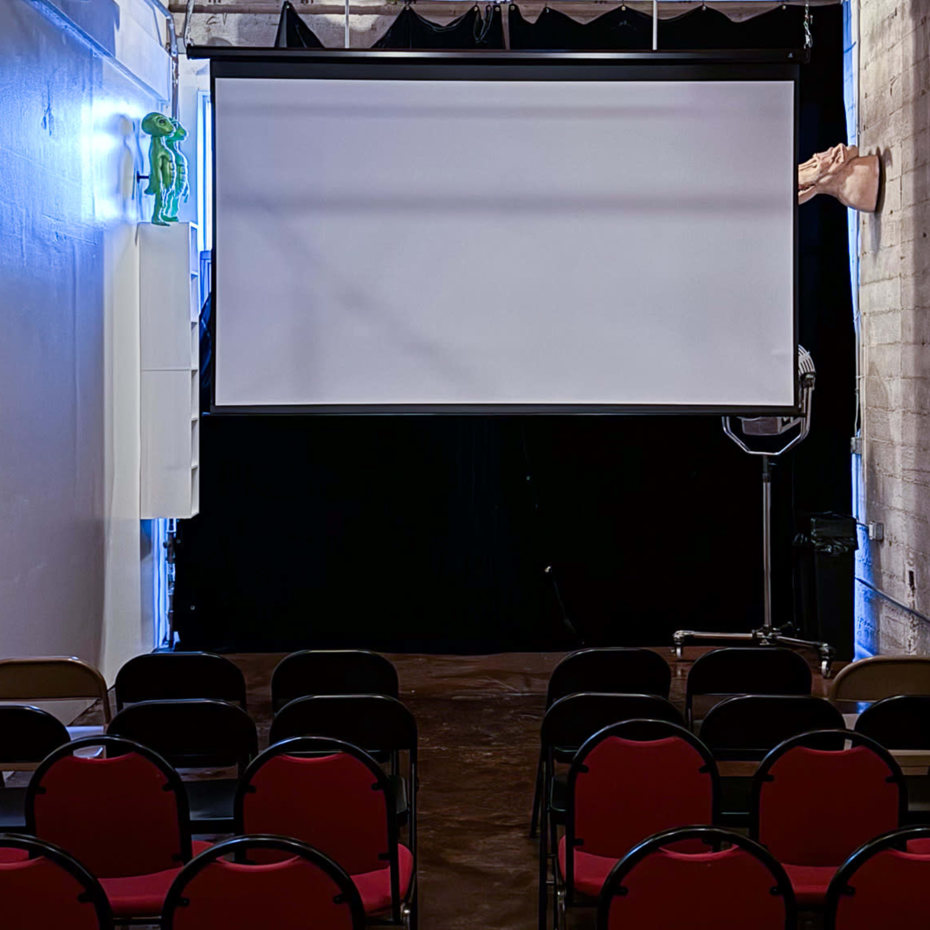 A small auditorium with red chairs arranged in rows facing a large blank projection screen, set against a dark backdrop.