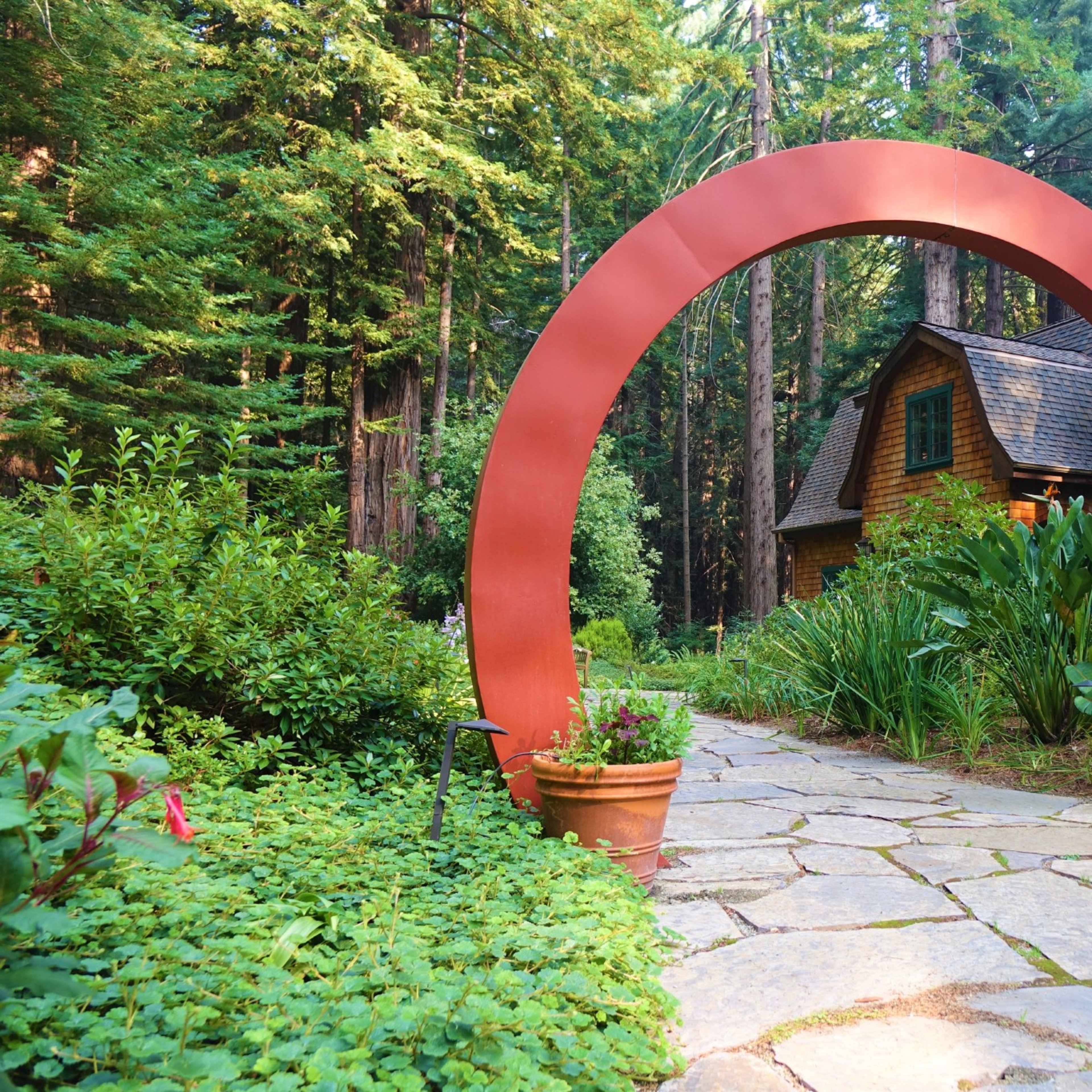 A red arch frames a stone pathway leading to a house nestled among tall trees and lush greenery.
