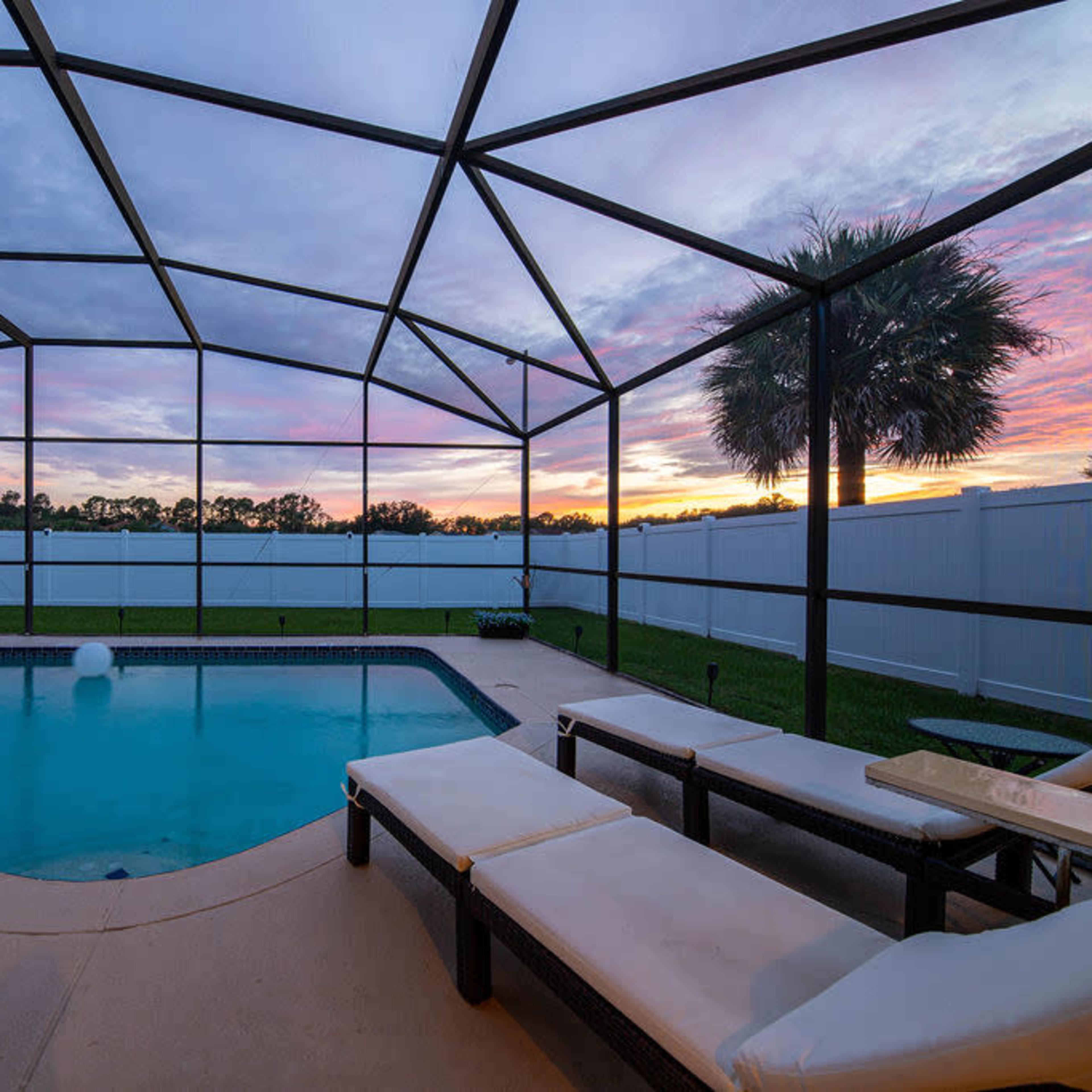A swimming pool enclosed by a screen with lounge chairs beside it, under a colorful sunset sky.