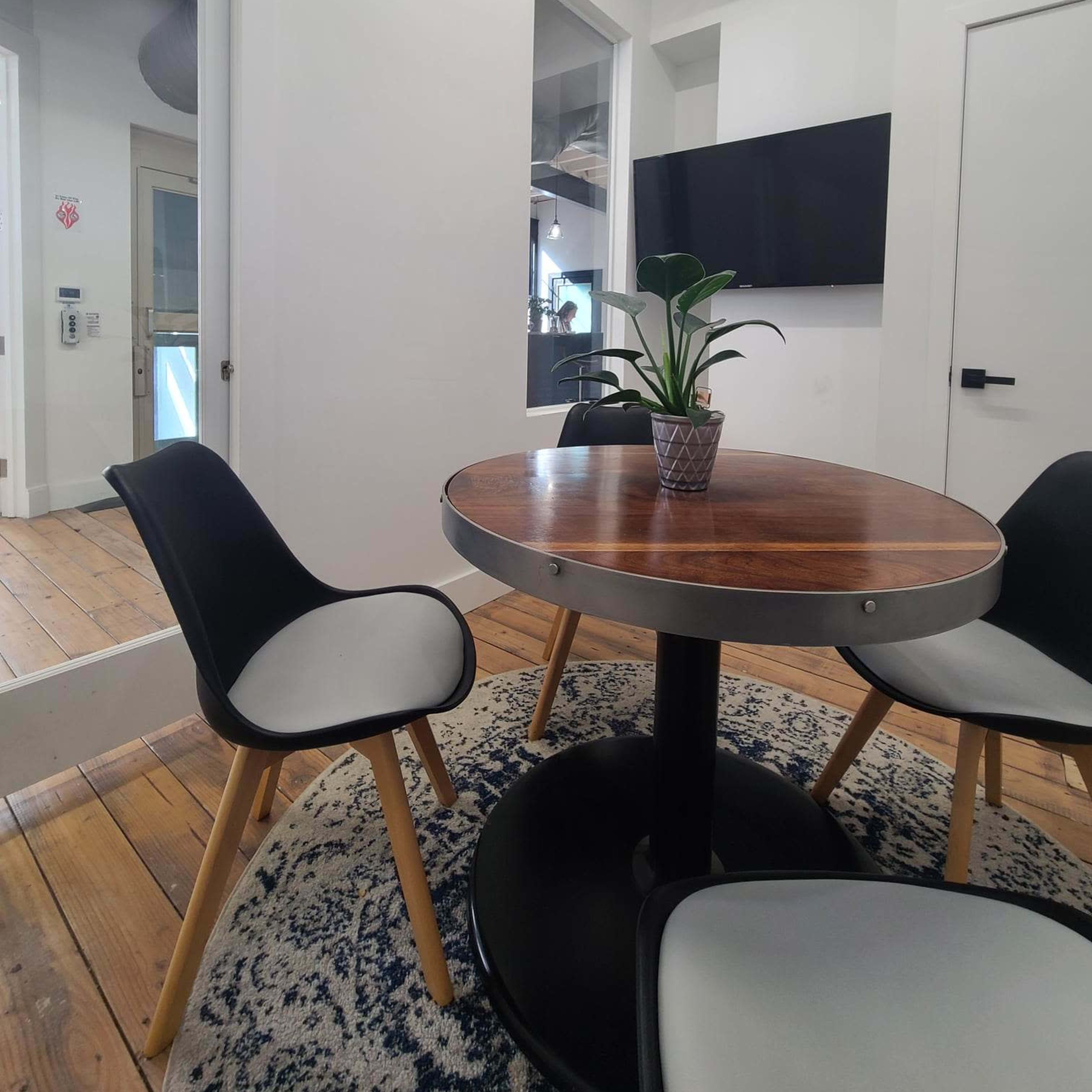 A small meeting room features a round wooden table and three black chairs with light-colored seats, accompanied by a plant in a pot.