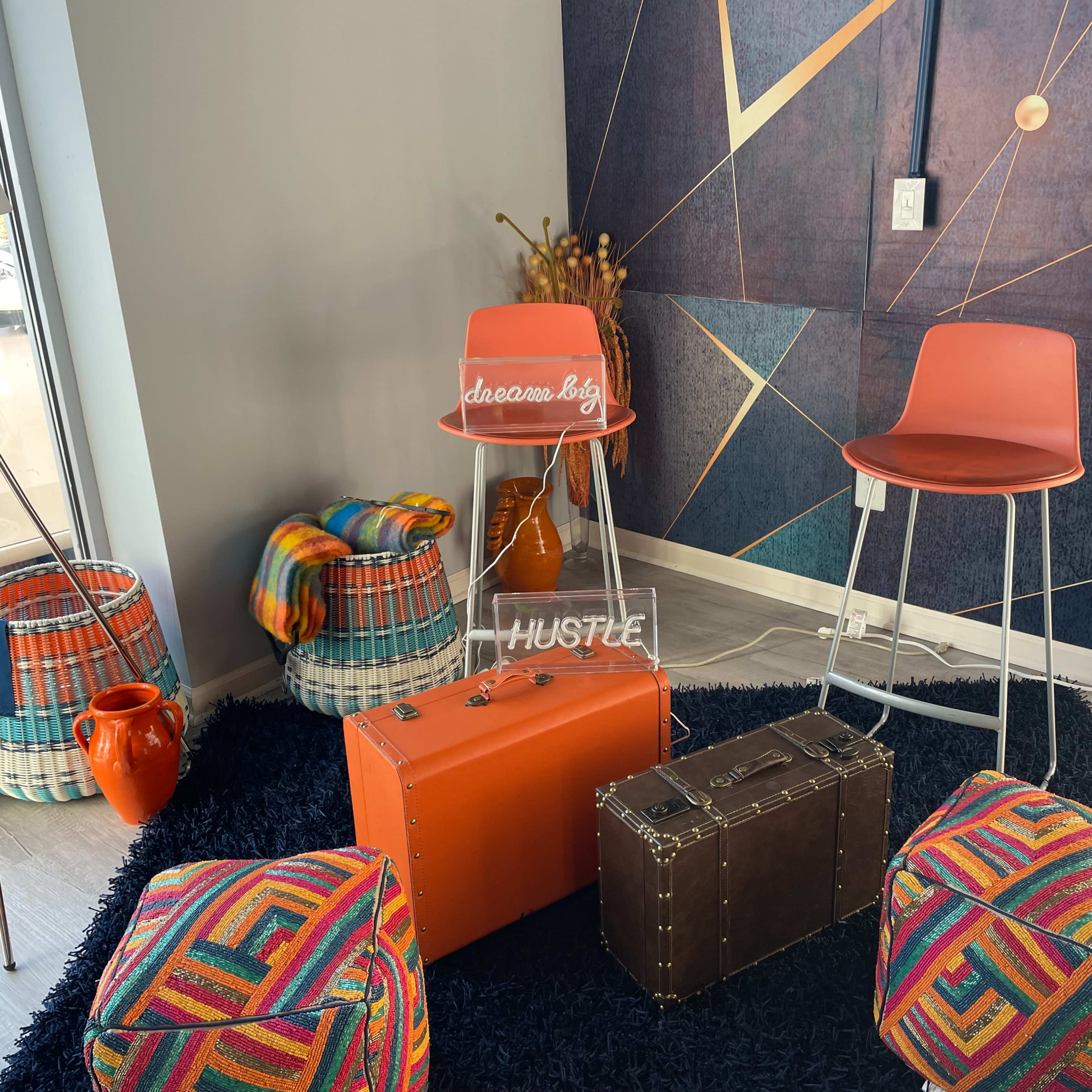 The scene features two orange bar stools, colorful poufs, a vintage-style trunk, and woven baskets against a geometric wall design.